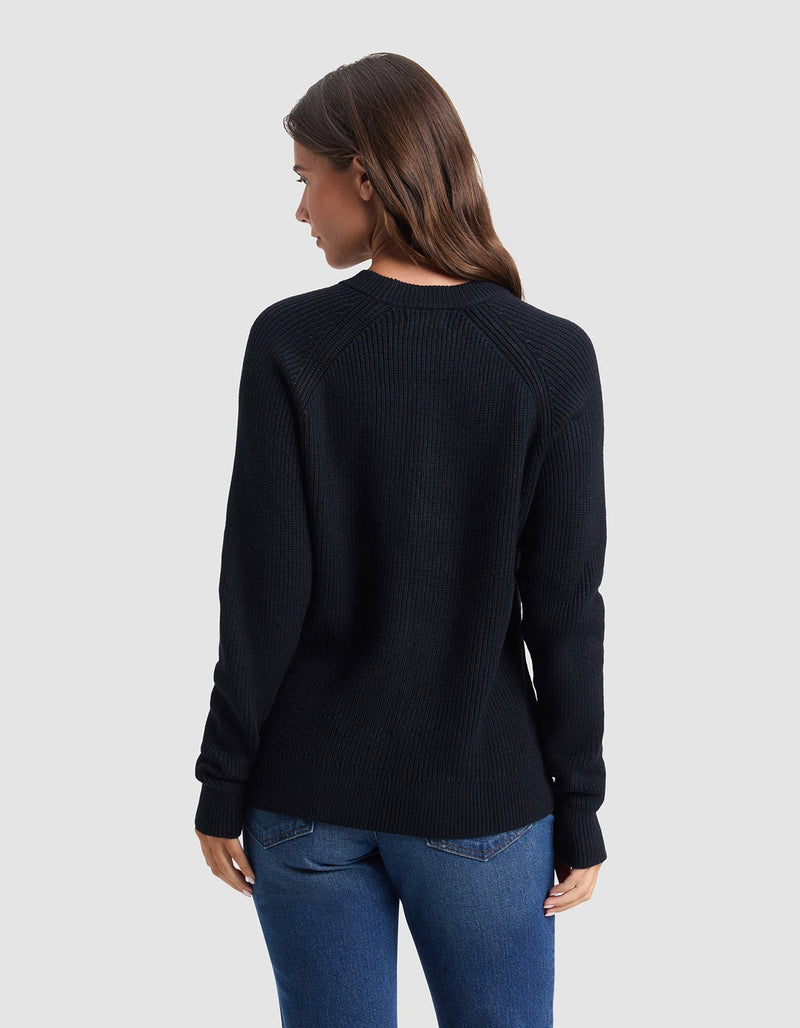 A woman with long brown hair, wearing the Cozy Earth Women's Classic Crewneck and blue jeans, stands facing away from the camera against a plain light gray background.
