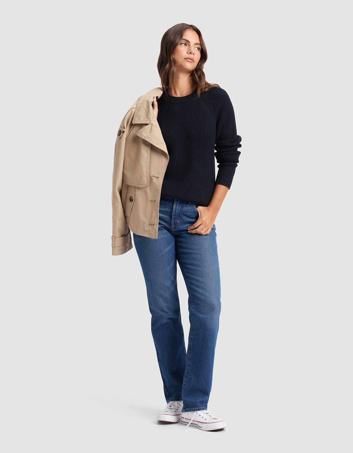 A woman stands against a plain background, wearing Cozy Earth's Women's Classic Crewneck in black, styled with blue jeans and white sneakers. She drapes a tan coat over one shoulder and glances to the side with one hand in her pocket.