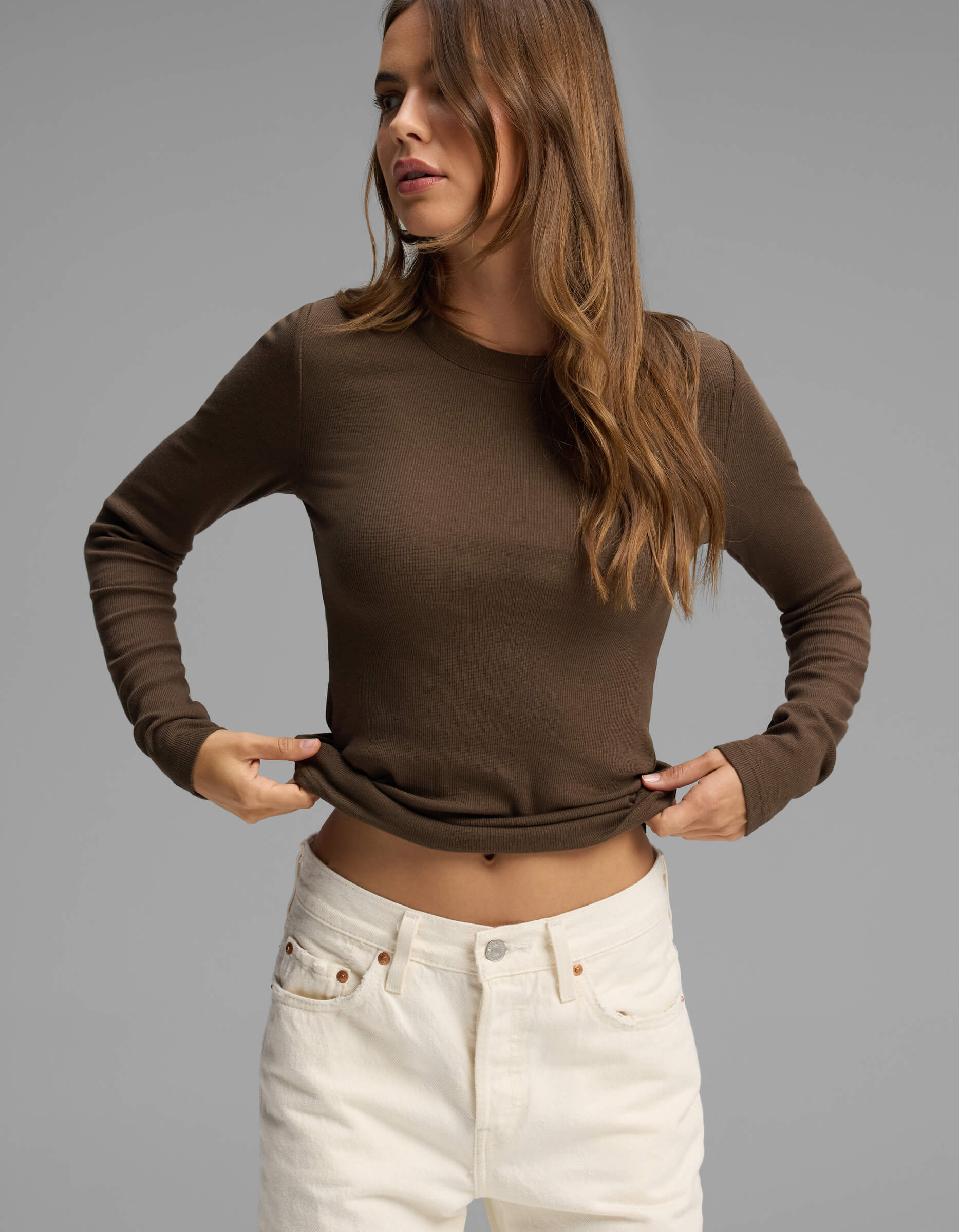 A woman with long brown hair wears the Cozy Earth Women's Fine Ribbed Long Sleeve Tee in brown, paired with high-waisted white jeans, standing against a plain gray background.|Color:Espresso