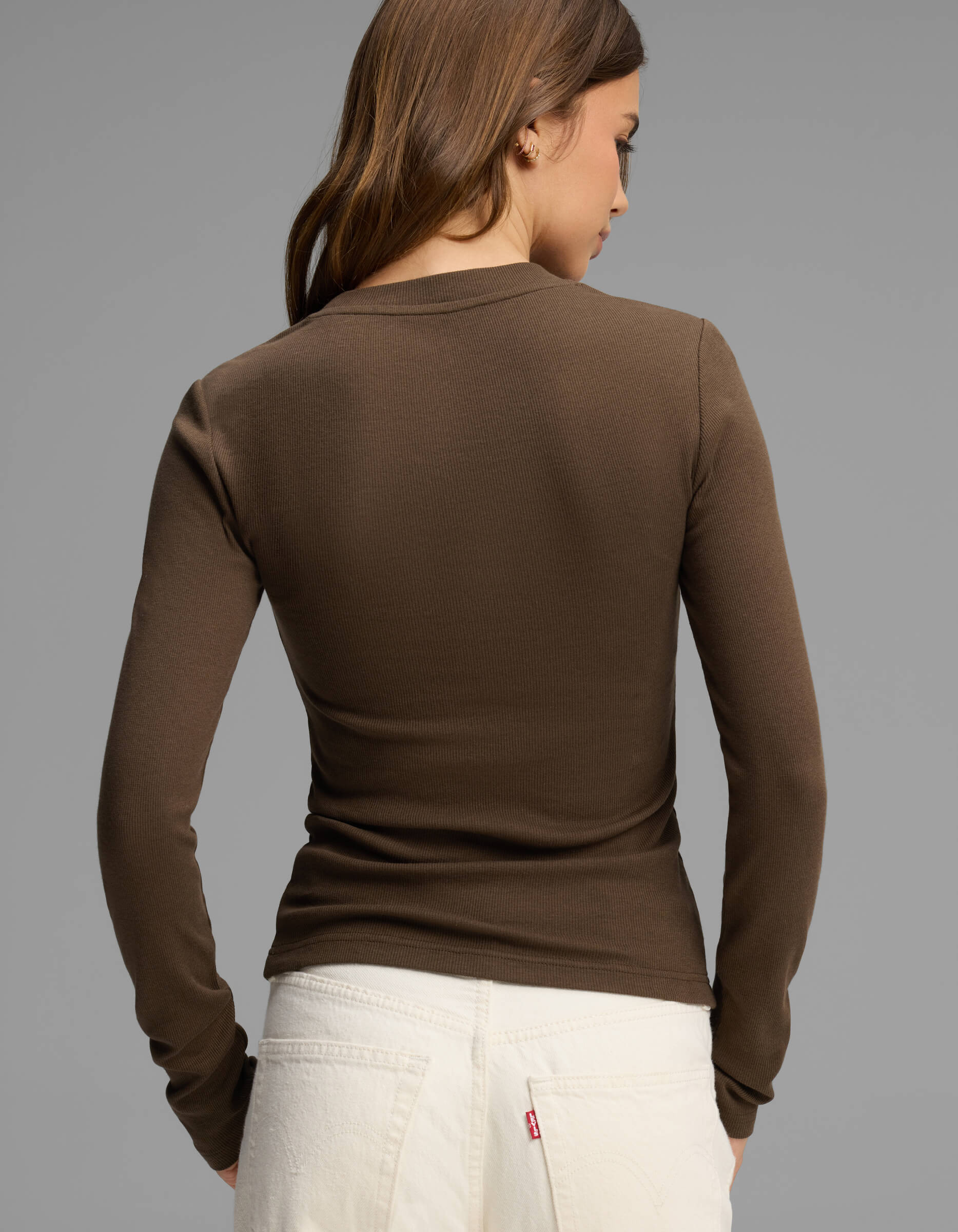 A woman with brown hair, wearing the Cozy Earth Women's Fine Ribbed Long Sleeve Tee in brown and white jeans, is standing with her back to the camera against a plain gray background.