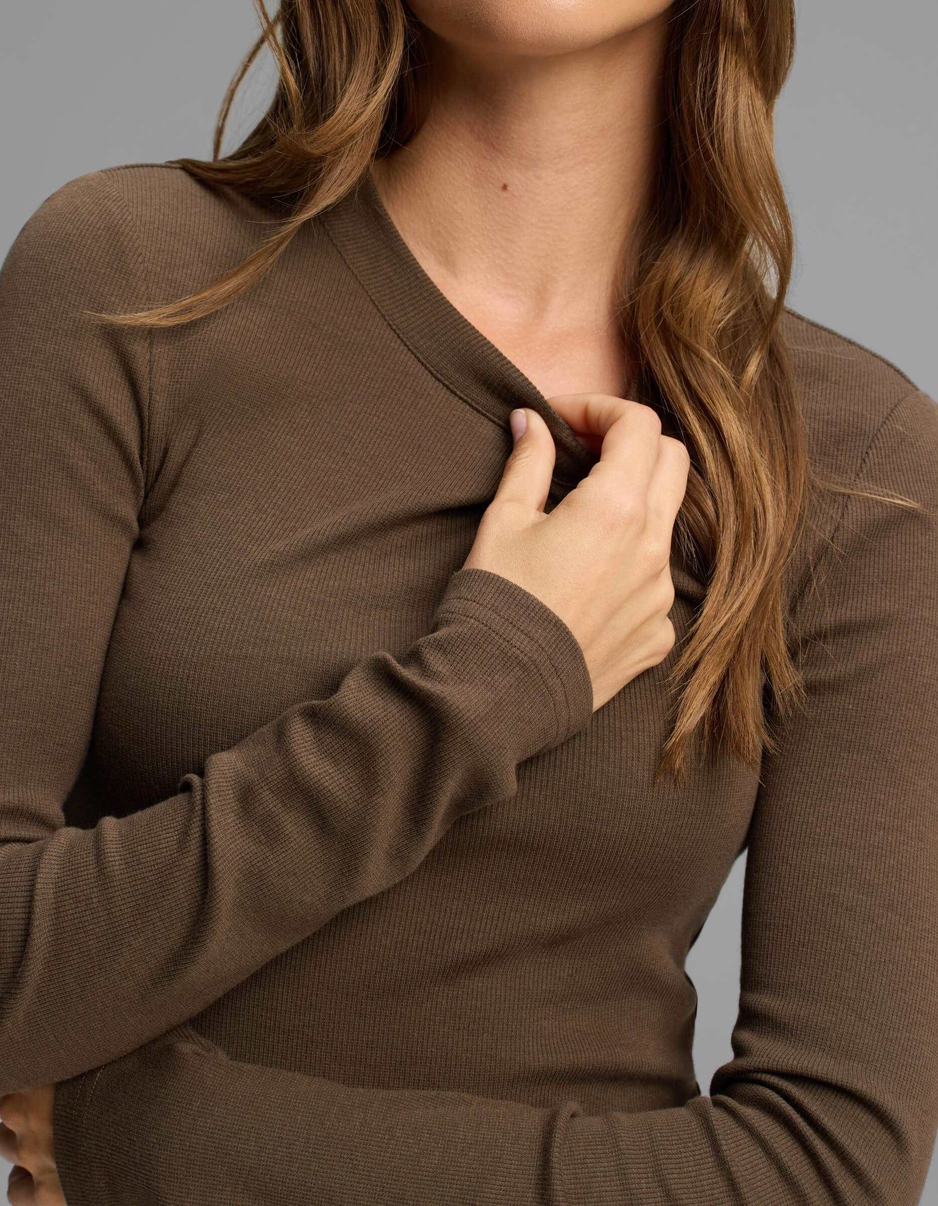 A person with long, wavy brown hair is wearing the Cozy Earth Women's Fine Ribbed Long Sleeve Tee in brown. They are holding the collar with one hand; their face is partially out of frame and the background is plain gray.