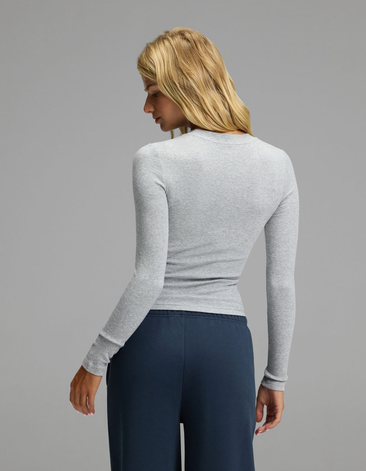A woman with long blonde hair, wearing the Cozy Earth Women’s Fine Ribbed Long Sleeve Tee in light gray and dark blue pants, stands facing away and looks over her shoulder against a plain gray background.|Color:Heather Grey