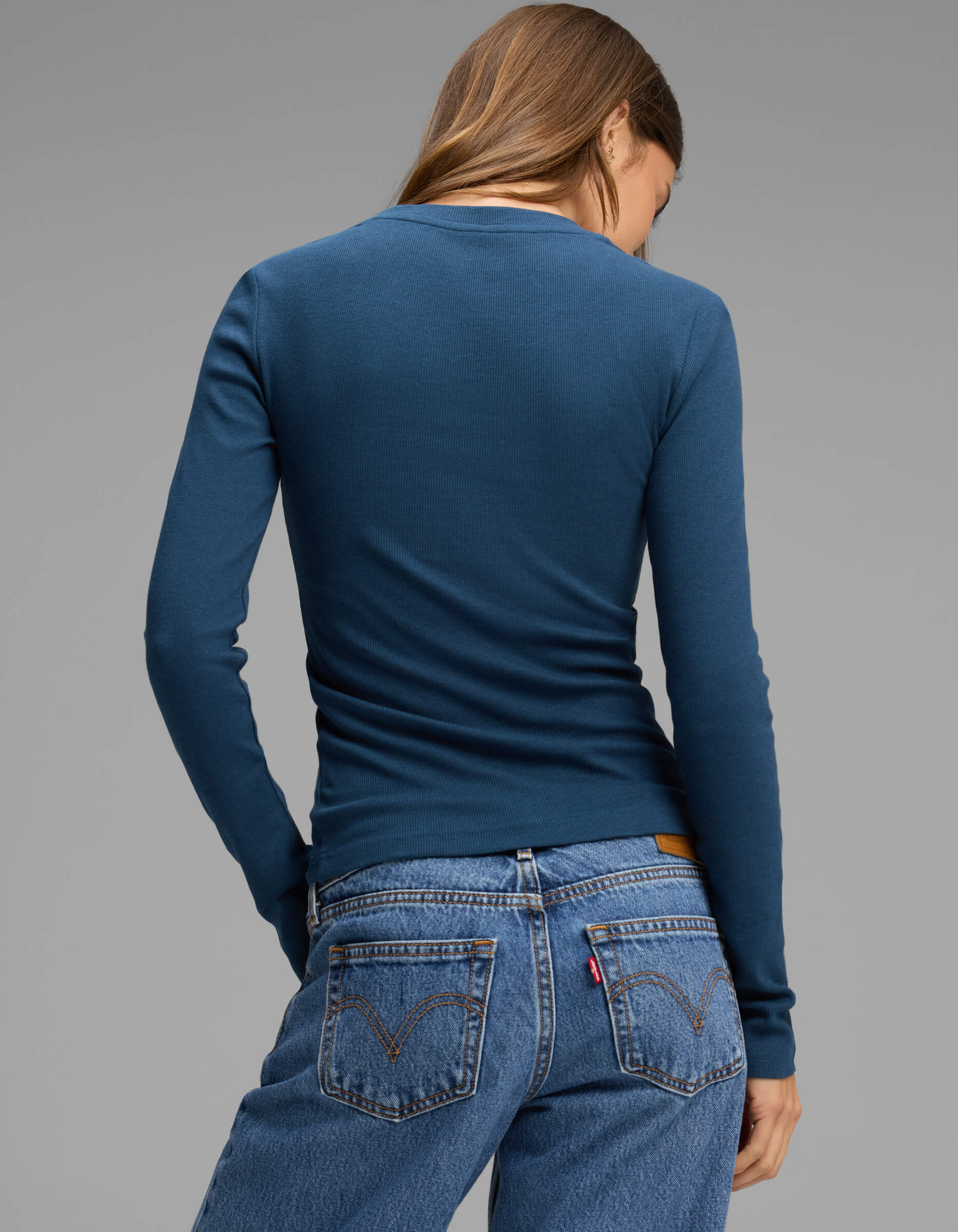 A woman with long brown hair, seen from behind against a plain gray background, is wearing the Cozy Earth Women's Fine Ribbed Long Sleeve Tee in blue along with blue jeans.