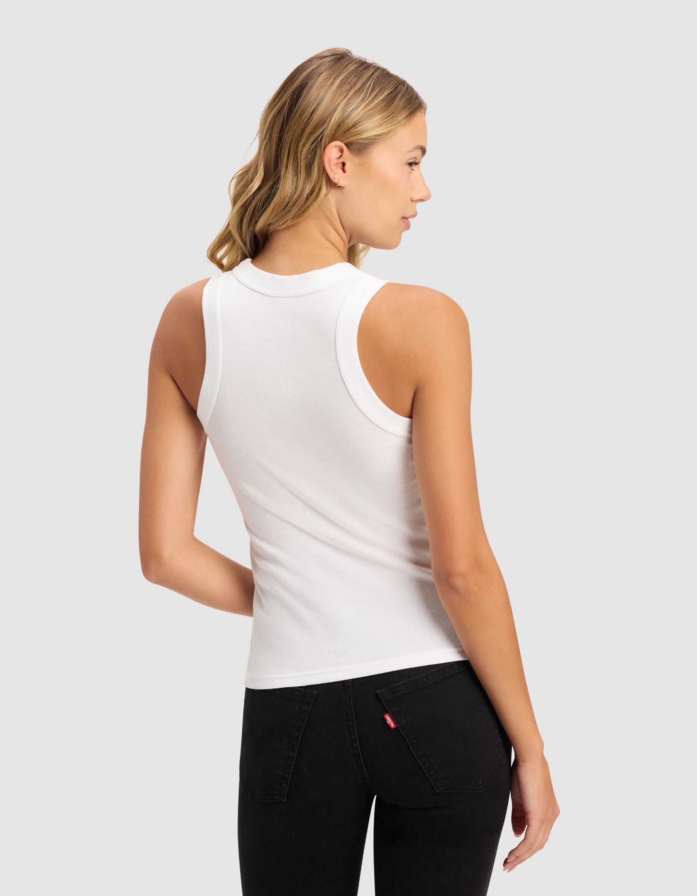 A woman with blonde wavy hair stands with her back to the camera, wearing Cozy Earth's Women's Fine Ribbed Tank in white and black jeans, set against a light gray background.