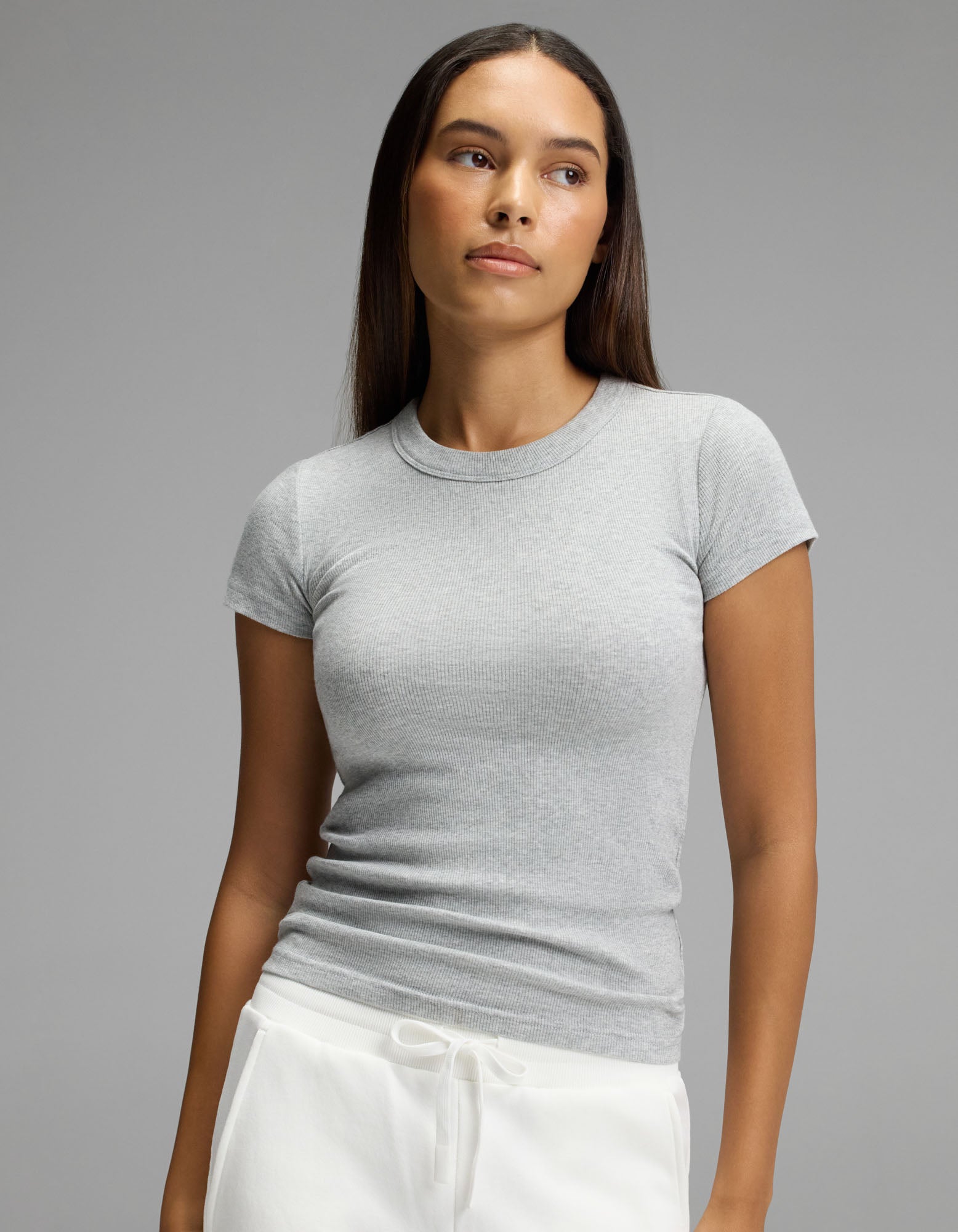 A woman with long brown hair wears the Cozy Earth Women’s Fine Ribbed Tee in light grey and white drawstring pants, standing against a plain grey background and looking slightly to her left with a neutral expression.