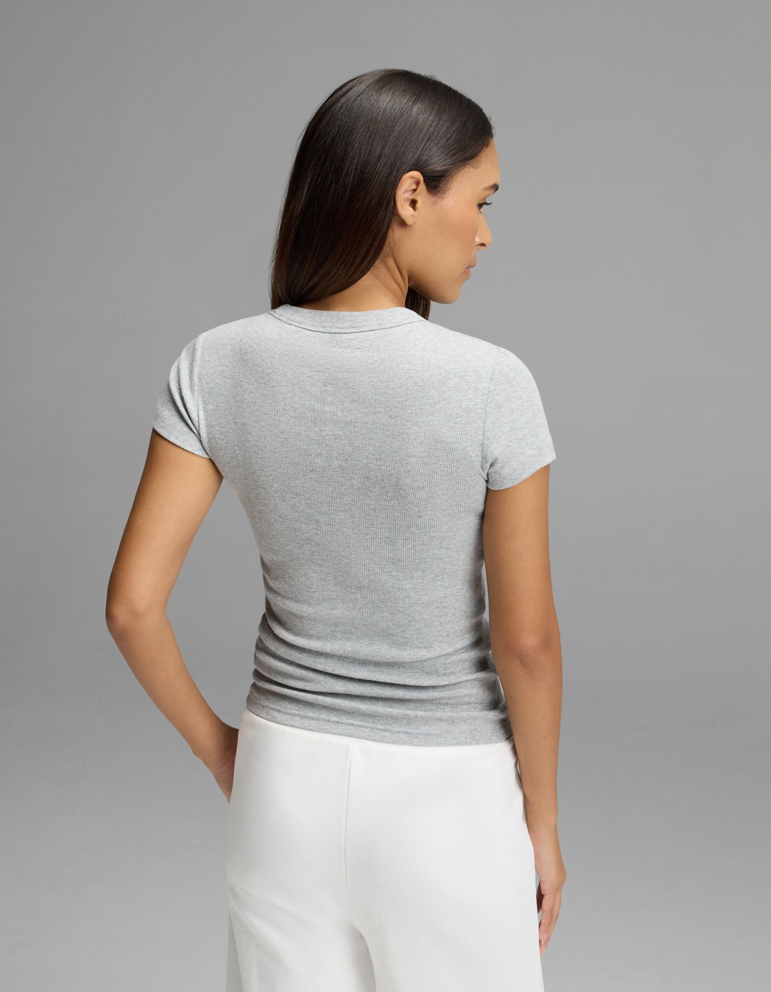 A woman with long dark hair is standing with her back to the camera, wearing the Cozy Earth Women’s Fine Ribbed Tee in gray and white pants against a plain gray background.