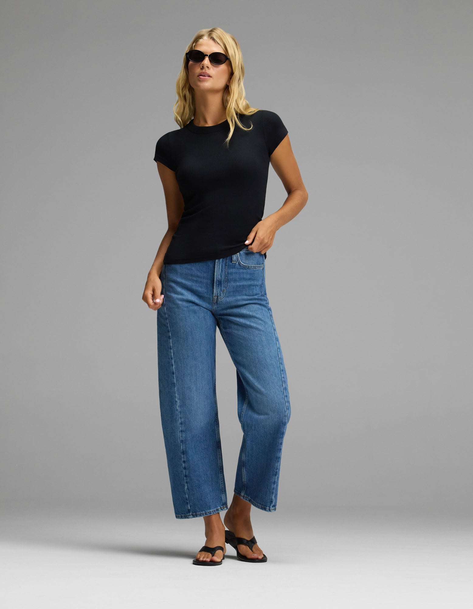 A woman stands confidently against a plain gray background, wearing sunglasses, blue wide-leg jeans, black sandals, and the Cozy Earth Women’s Fine Ribbed Tee.