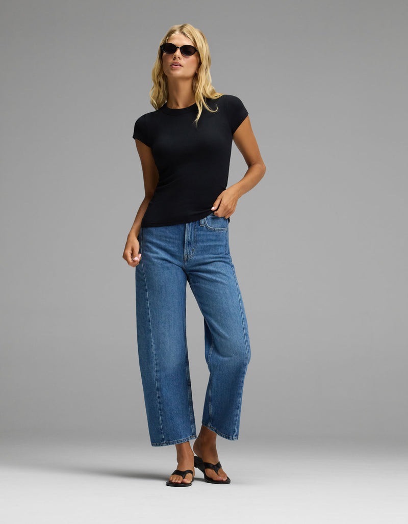A woman stands confidently against a plain gray background, wearing sunglasses, blue wide-leg jeans, black sandals, and the Cozy Earth Women’s Fine Ribbed Tee.