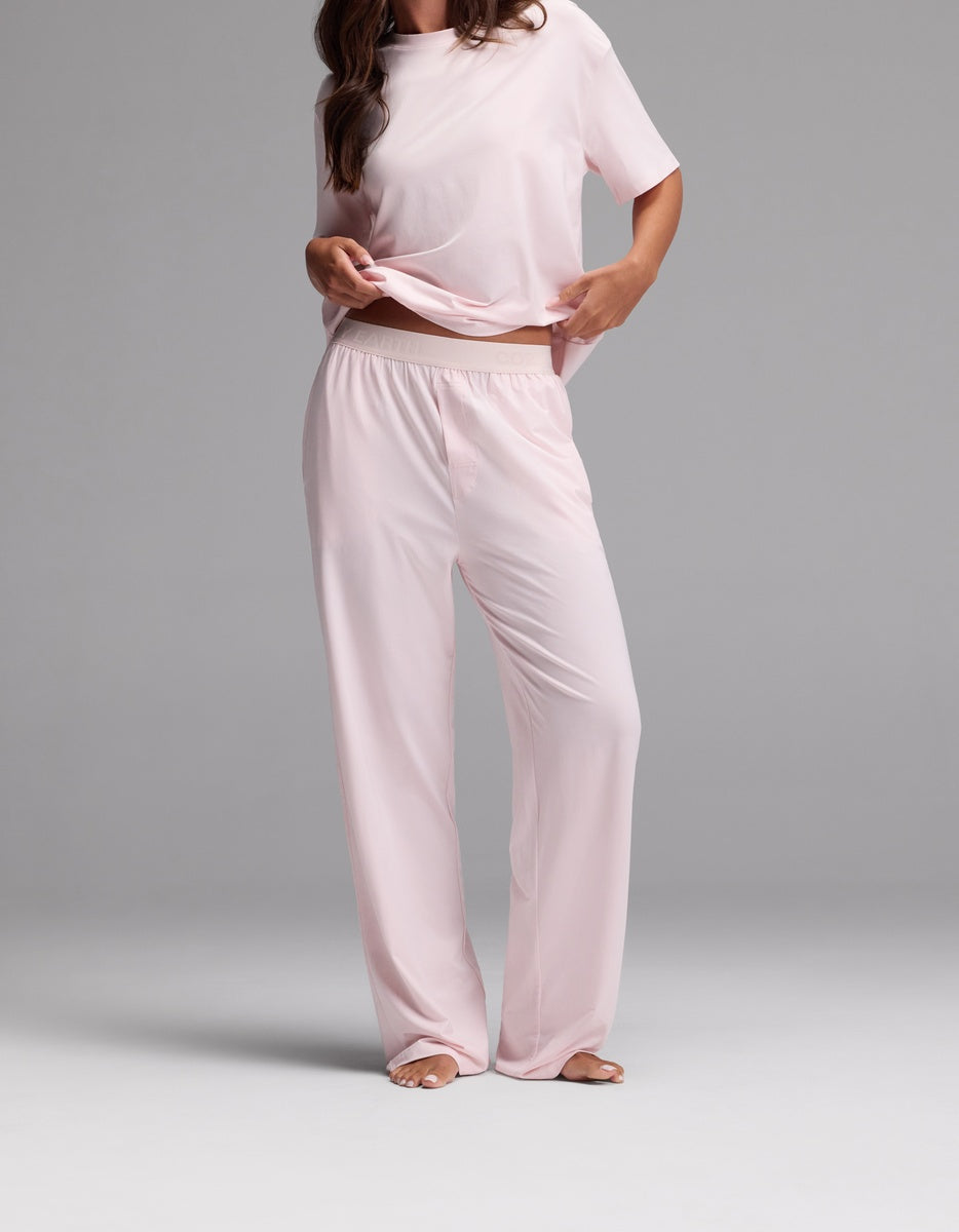A woman stands barefoot wearing the Cozy Earth Women’s First Layer Pajama Pant in Ballet, a loose-fitting, light pink short-sleeve pajama set with wide-leg pants. She holds up part of her shirt, and her head is not visible in the image.