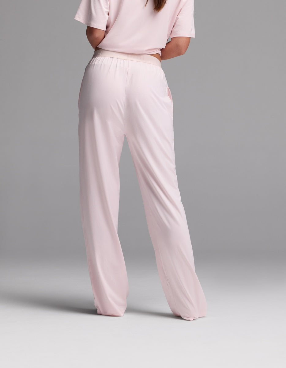 A woman is shown from the back, standing against a plain gray background, wearing the Cozy Earth Women’s First Layer Pajama Pant in light pink with a matching short-sleeve top.