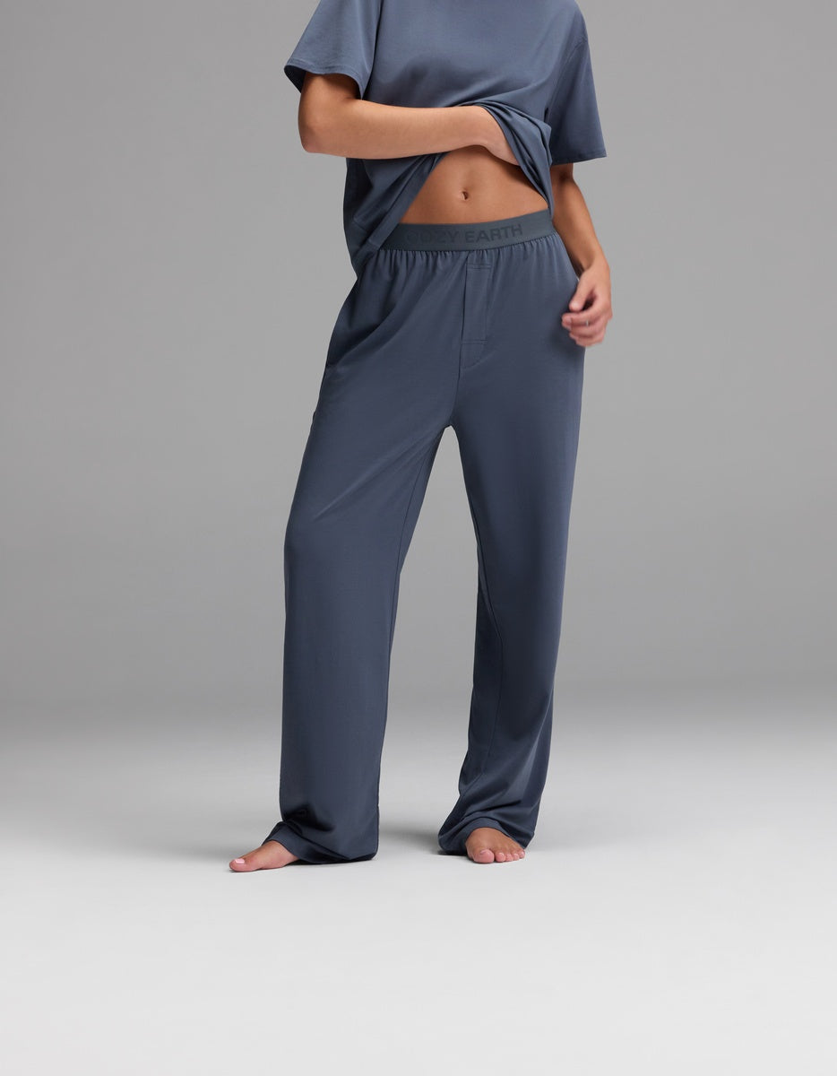 A person stands barefoot on a plain gray background, wearing Cozy Earth Women’s First Layer Pajama Pant in Dawn with a matching T-shirt, holding up part of the shirt to reveal the pajama pant’s waistband.|Color:Dawn