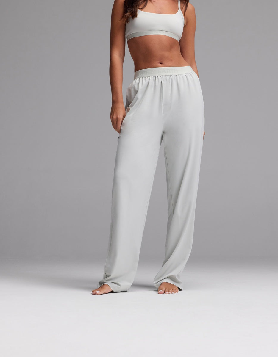 A woman wearing Cozy Earth’s Women’s First Layer Pajama Pant in Laurel stands barefoot against a plain gray background, with her hands in her pockets and her head cropped out of the image.