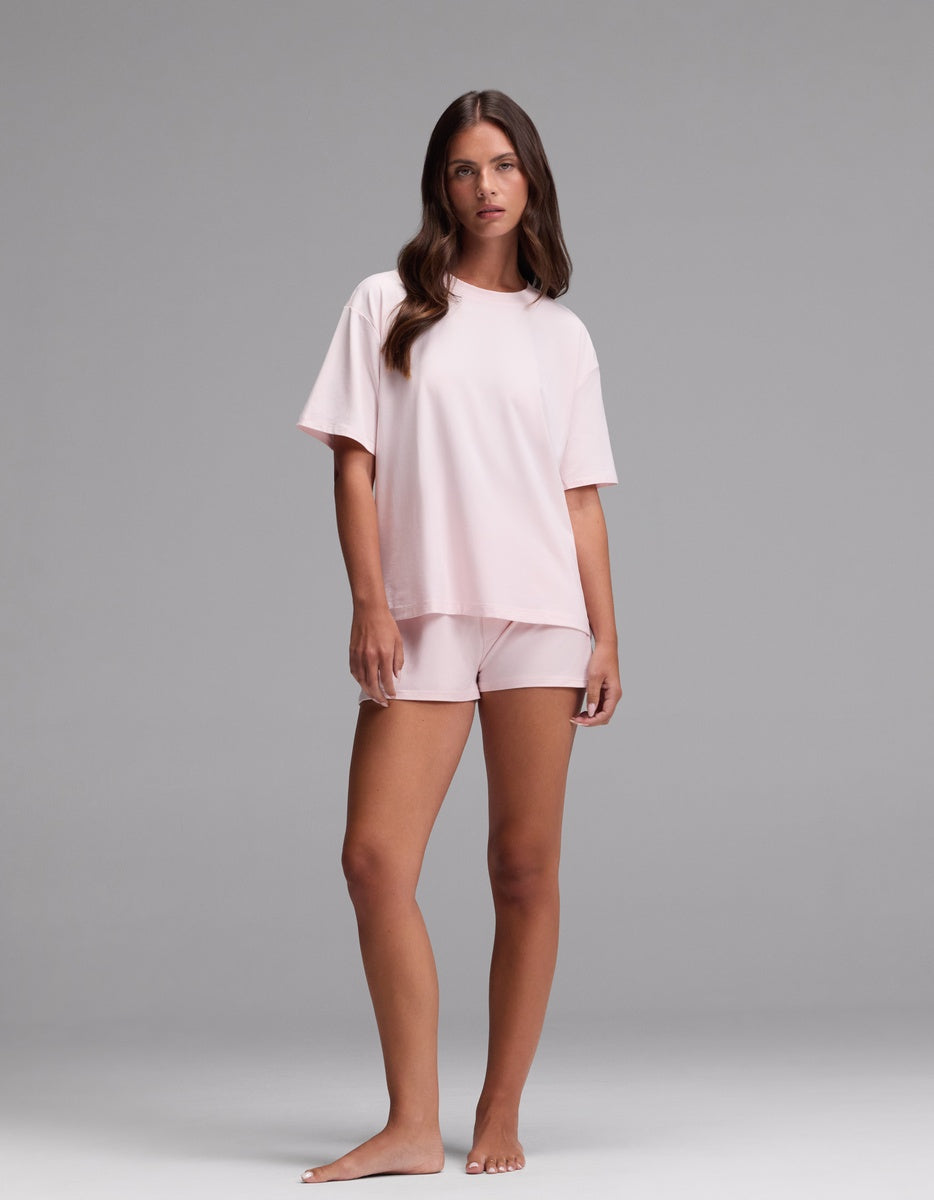 A woman stands barefoot against a plain background, wearing the Cozy Earth Women’s First Layer Pajama Short Sleeve Tee & Boxer Short Set in Ballet. She has long brown hair and a neutral expression.|Color:Ballet