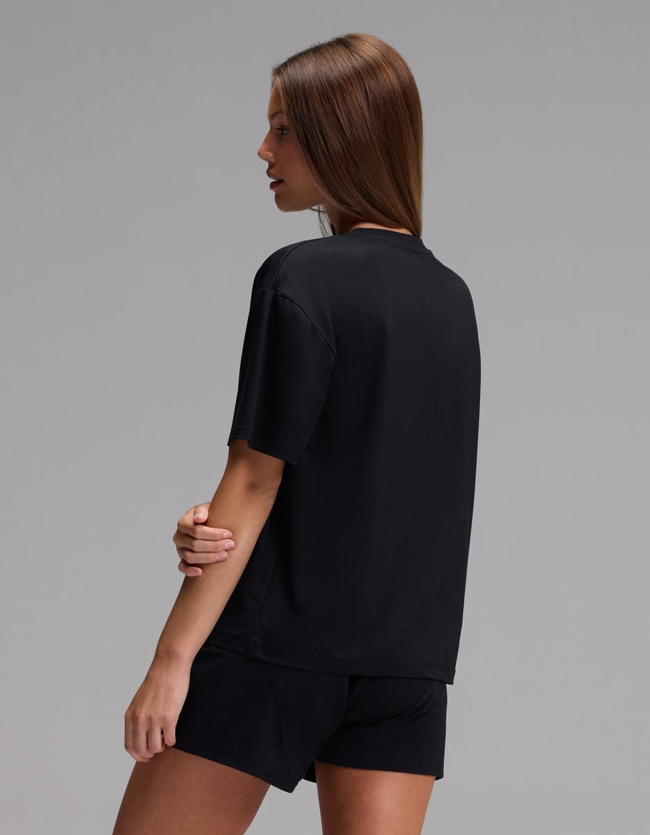 A person with straight brown hair is wearing the Cozy Earth Women’s First Layer Pajama Short Sleeve Tee & Boxer Short Set in black, standing sideways against a plain gray background.