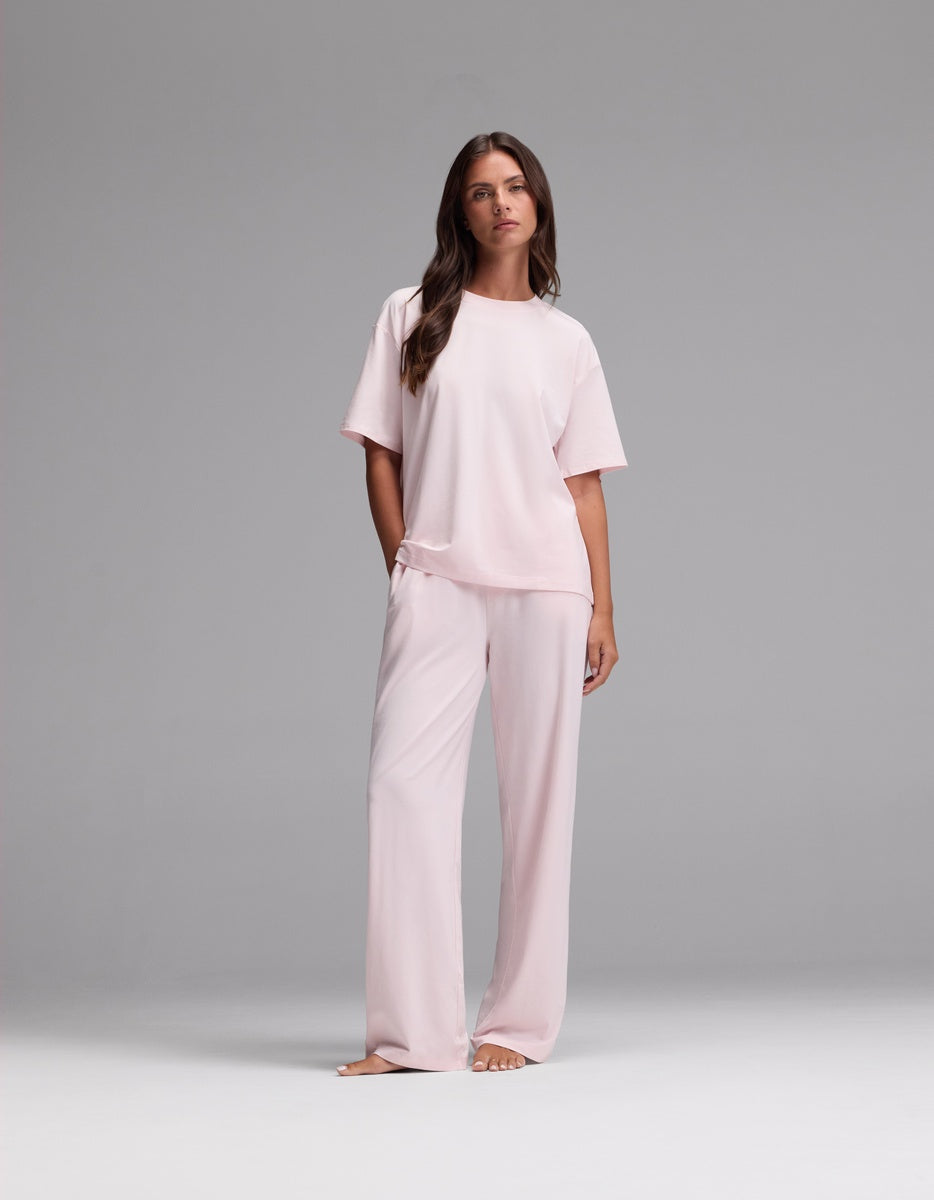A woman stands barefoot against a plain gray background, wearing the Cozy Earth Women’s First Layer Pajama Short Sleeve Tee & Pajama Pant Set in Ballet—a loose-fitting, light pink ensemble featuring short sleeves and wide-leg pants.|Color:Ballet