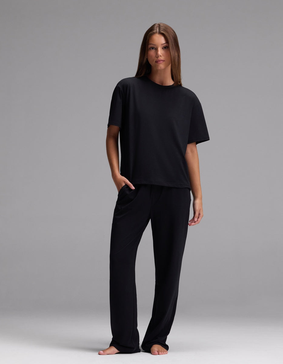 A woman stands barefoot against a plain background, wearing the Cozy Earth Women’s First Layer Pajama Short Sleeve Tee & Pajama Pant Set in Jet Black, with her left hand in her pocket.|Color:Jet Black