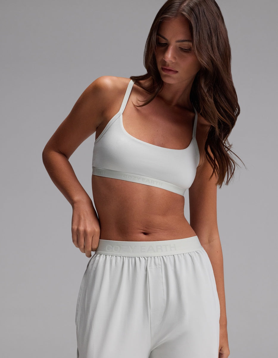 A woman with long brown hair wears the Cozy Earth Women’s First Layer Soft Bra in Laurel, paired with matching sweatpants, looking down against a plain gray background.