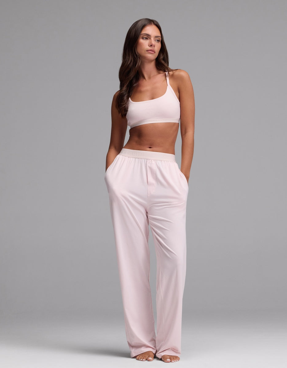 A woman stands with her hands in her pockets against a plain gray background, wearing the Cozy Earth Women’s First Layer Pajama Soft Bra & Pajama Pant Set in Ballet.|Color:Ballet