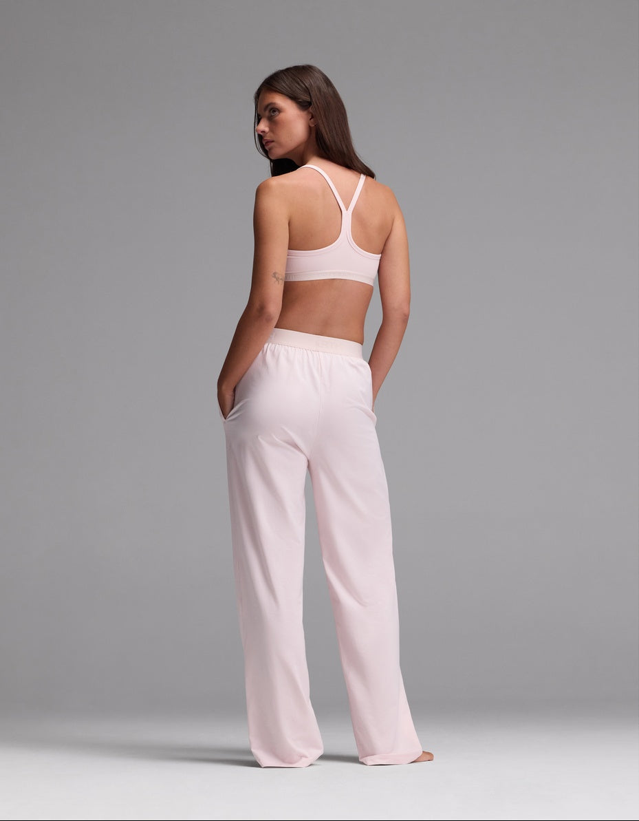 A woman stands with her back to the camera, wearing the Cozy Earth Women’s First Layer Pajama Soft Bra & Pajama Pant Set in light pink. Her hands rest in her pockets as she looks over her shoulder against a plain gray background.|Color:Ballet