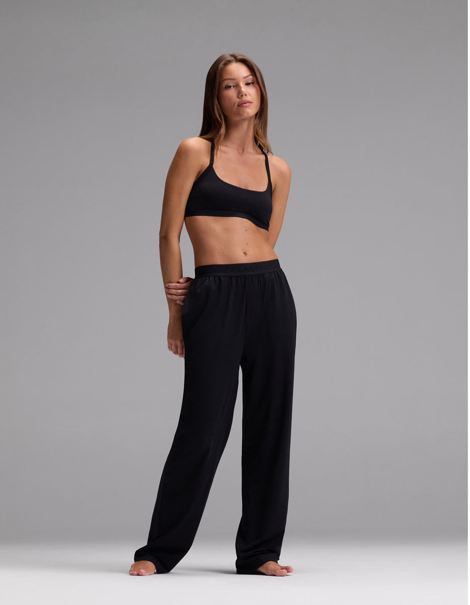 A woman stands barefoot against a gray background, wearing the Cozy Earth Women’s First Layer Pajama Soft Bra & Pajama Pant Set in Jet Black, with one hand at her side and the other bent at her elbow.|Color:Jet Black