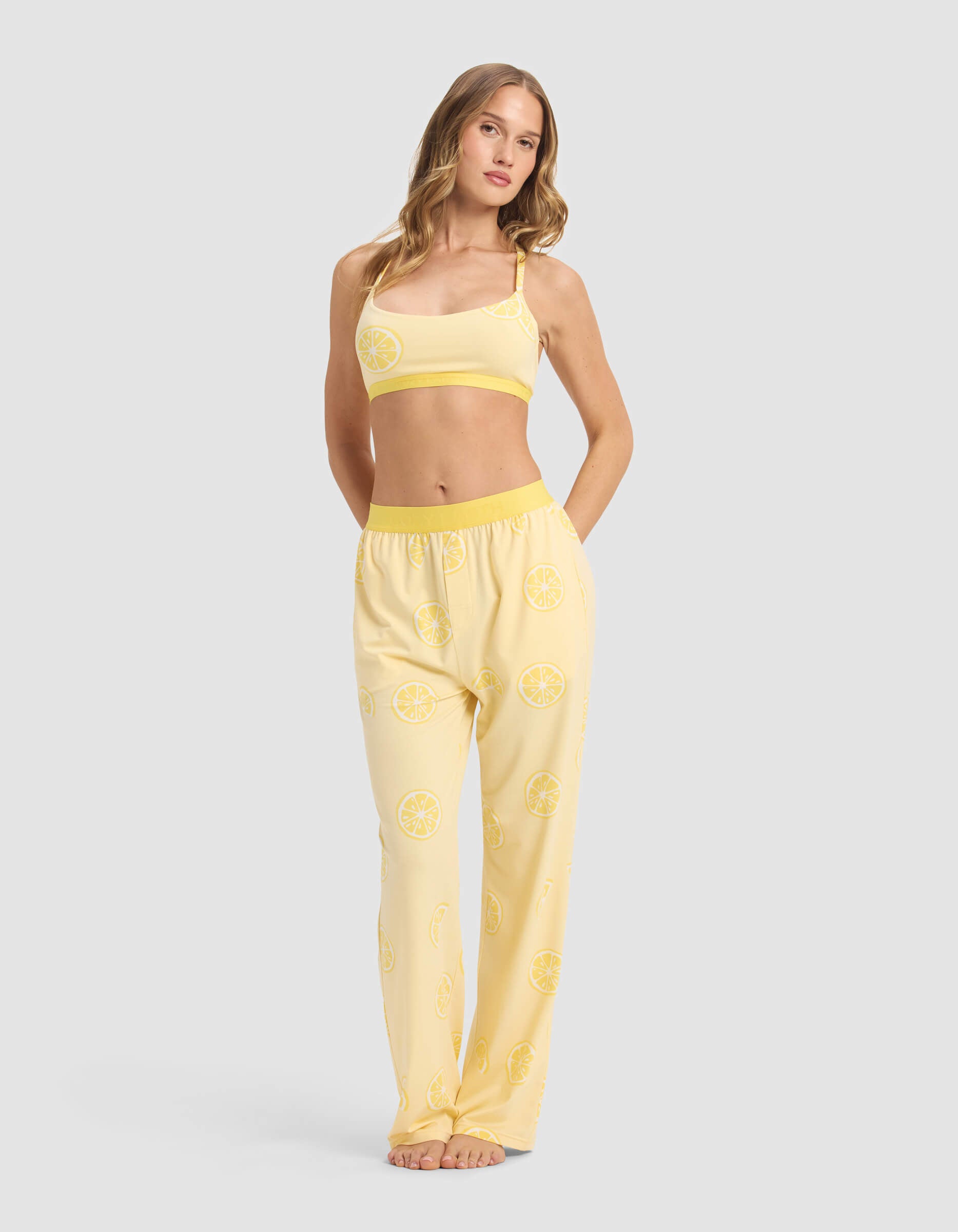 A woman with long light brown hair poses barefoot against a plain background, wearing Cozy Earth's Women's First Layer Pajama Soft Bra & Pant Set—a yellow set with a lemon slice pattern, featuring a crop top and loose pants with pockets.