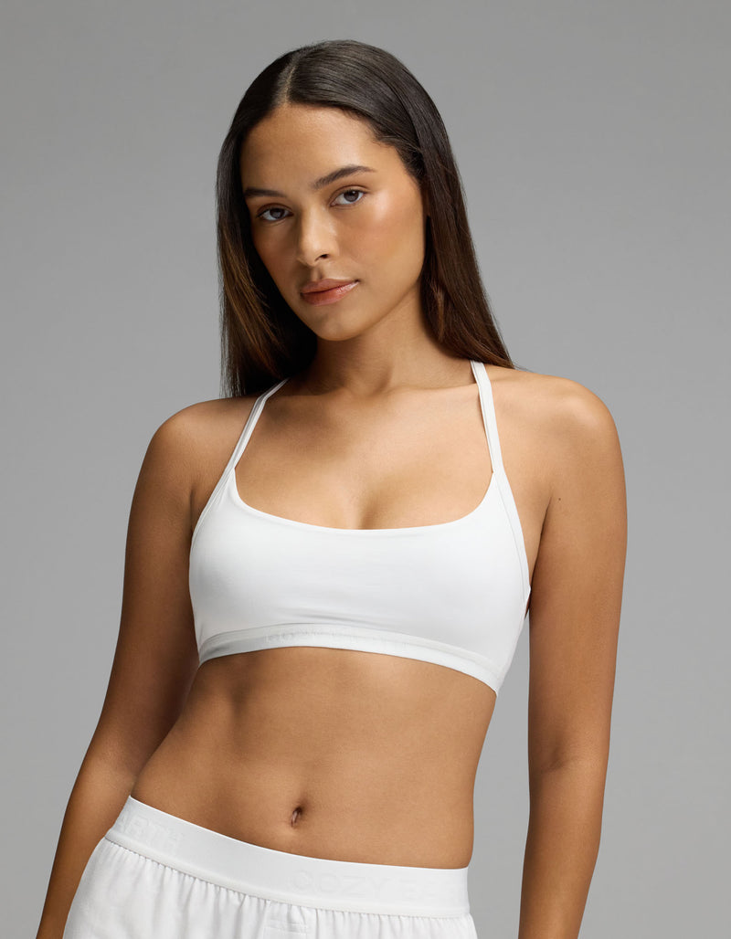 A woman with long brown hair stands confidently against a plain gray background, wearing Cozy Earth’s Women’s First Layer Soft Bra in white paired with white pants.