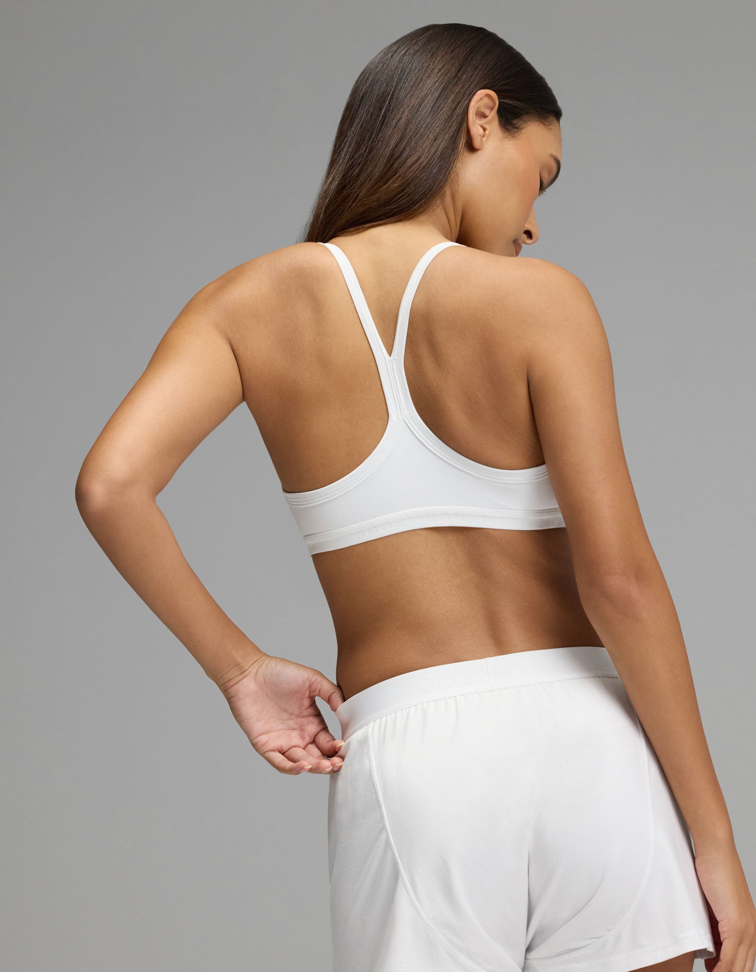 A woman stands with her back to the camera, wearing the Cozy Earth Women’s First Layer Soft Bra in white, paired with matching white shorts, against a plain gray background.