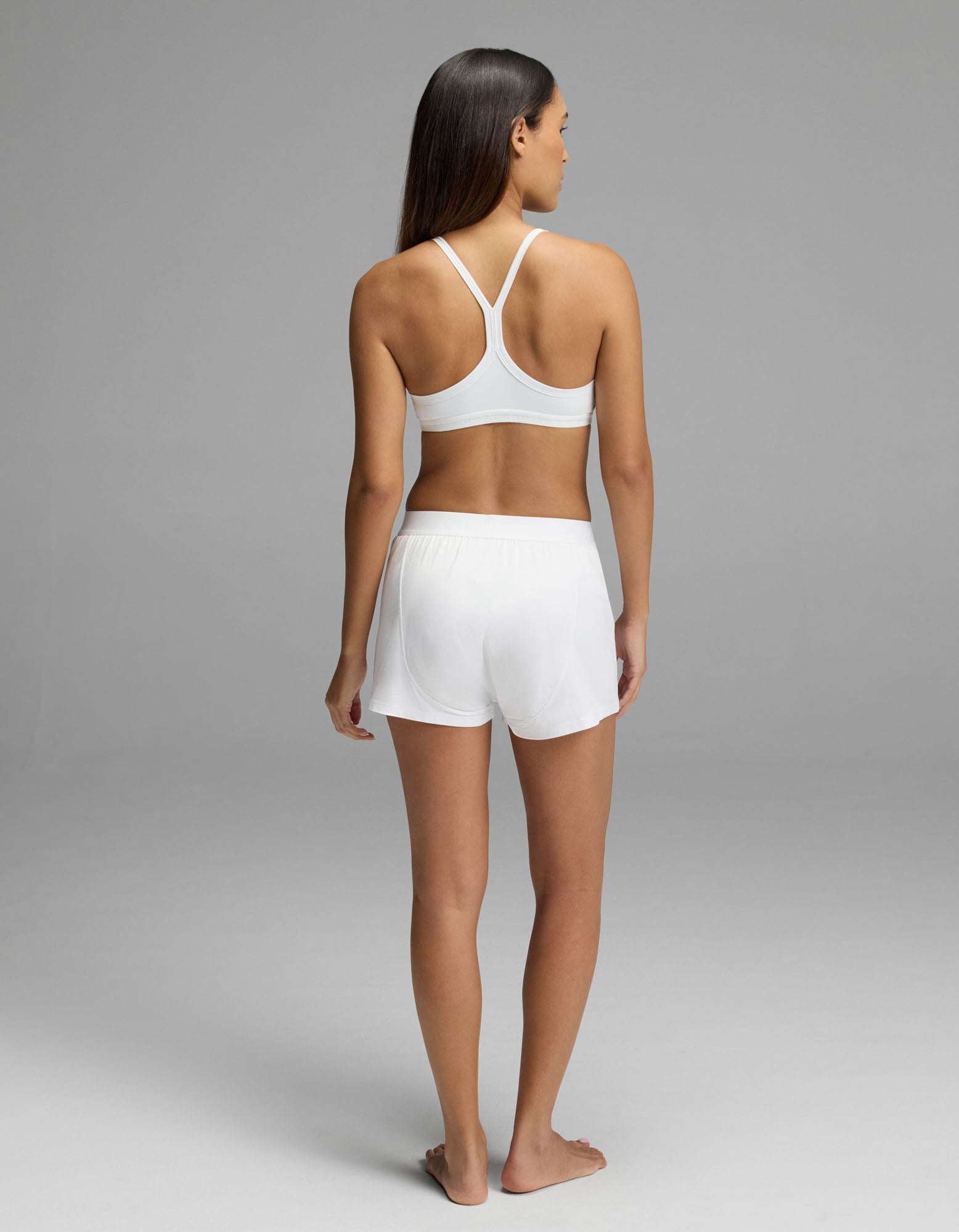 A woman stands barefoot on a gray background, facing away. She wears the Cozy Earth Women’s First Layer Pajama Soft Bra with a racerback design and matching Boxer Short Set in white.