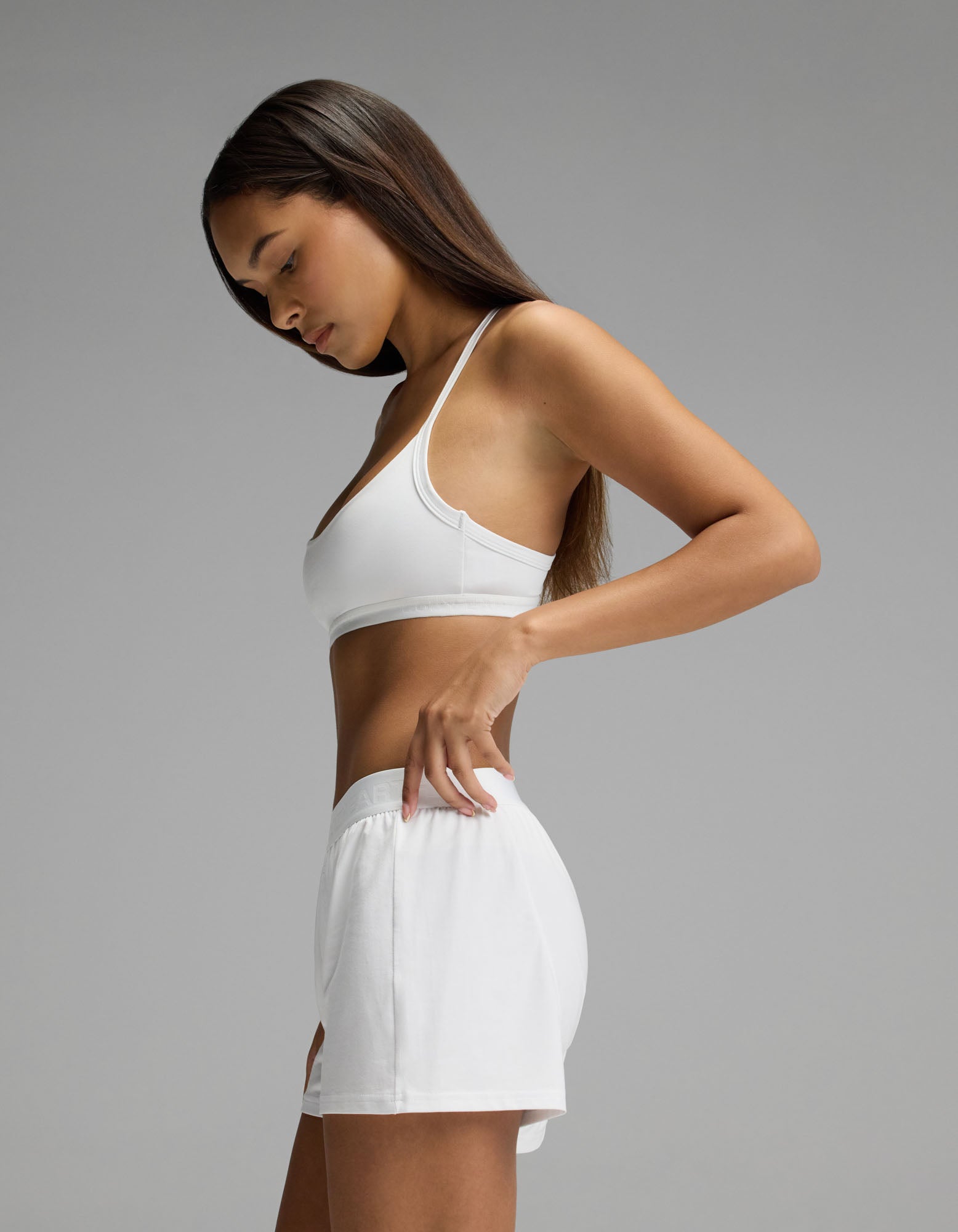 A woman wearing the Cozy Earth Women’s First Layer Pajama Soft Bra & Boxer Short Set in white stands in profile against a plain gray background, looking down and holding the waistband of her shorts.
