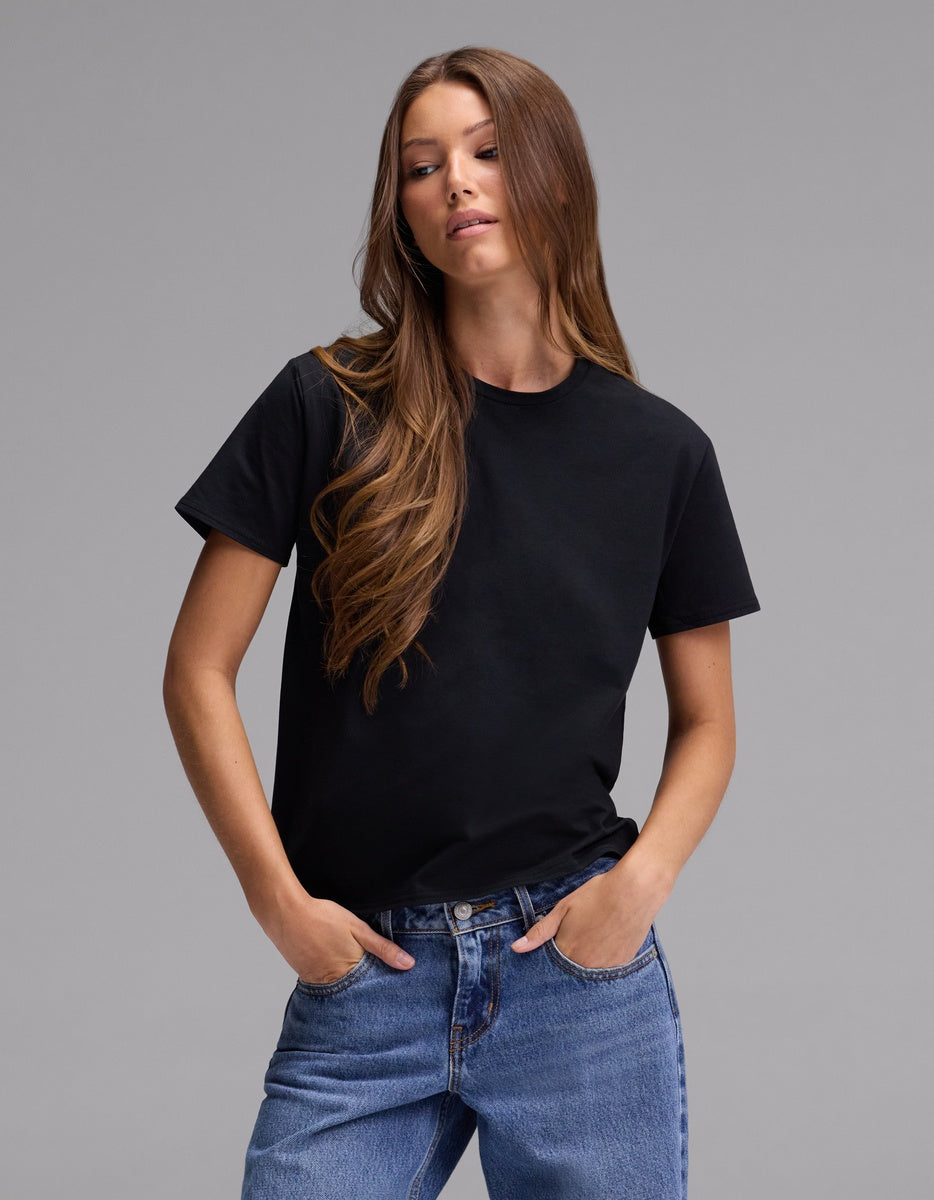 A woman with long brown hair wears the Cozy Earth Women’s Go-To Tee in black and blue jeans, standing with her hands in her pockets against a plain gray background.|Color:Jet Black