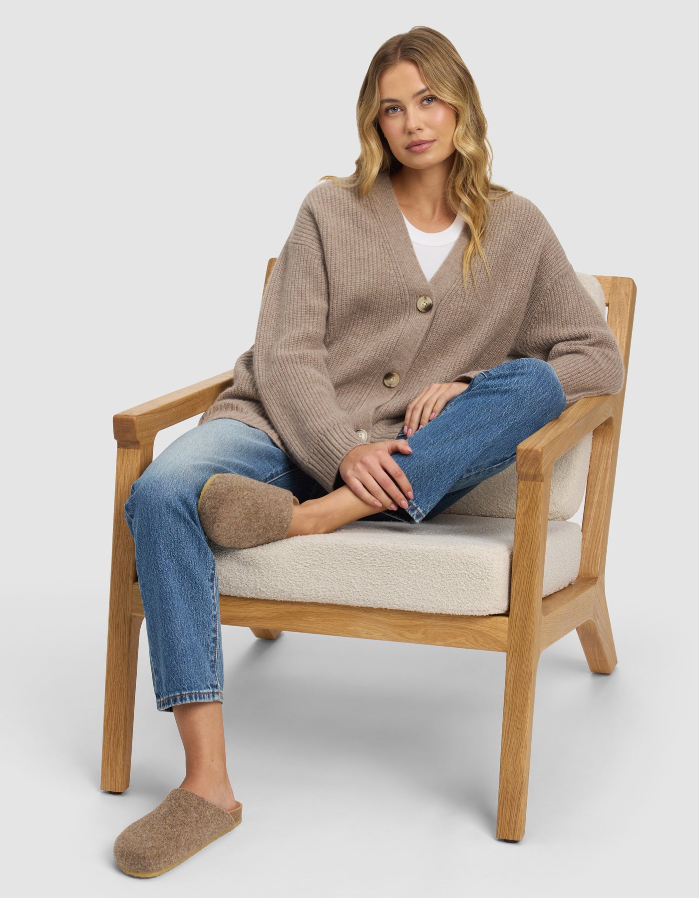A woman with long blonde hair sits relaxed on a wooden armchair, wearing blue jeans and Cozy Earth’s Lakehouse Clog slippers. One leg is crossed over the other, and she looks calmly at the camera.|Color:Chestnut