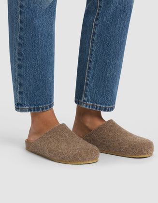 A person wearing blue jeans and Cozy Earth Lakehouse Clog slippers stands on a white background, with only their lower legs and feet visible.|Color:Chestnut