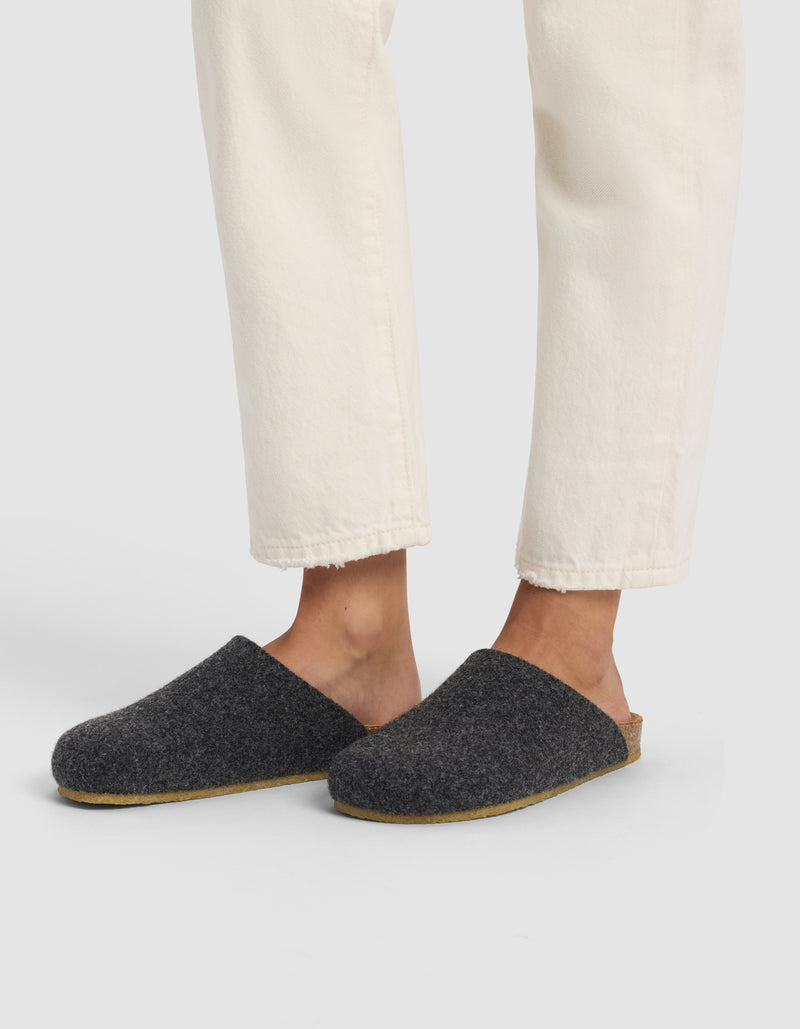 A person wearing off-white pants and Cozy Earth’s Lakehouse Clog—dark gray wool slippers with light brown soles—is standing against a plain light background.