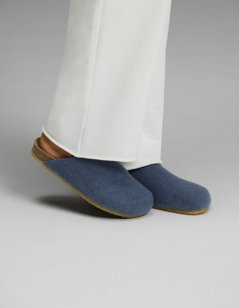 A person wearing white pants and Cozy Earth Lakehouse Clog slippers in light blue, featuring cork soles, stands on a light gray surface.