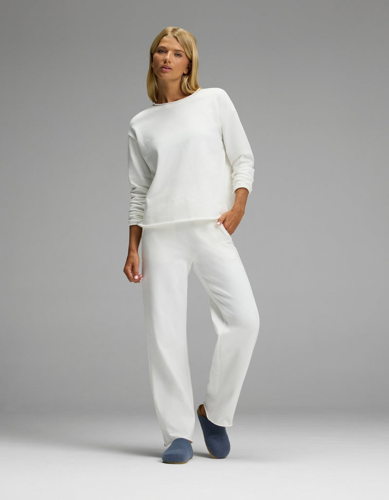 A woman stands in a relaxed pose wearing an all-white long-sleeve top and matching pants, with one hand in her pocket, against a plain gray background. She completes the look with Cozy Earth’s Lakehouse Clog slippers.