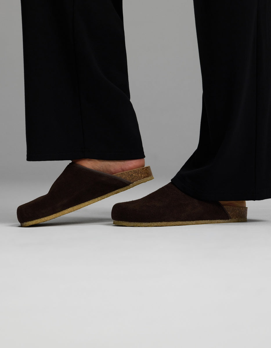 A person wearing black pants and the Cozy Earth Lakehouse Clog in Suede—dark brown slip-on clogs with cork soles—stands on a light, neutral background, with only their lower legs and feet visible.