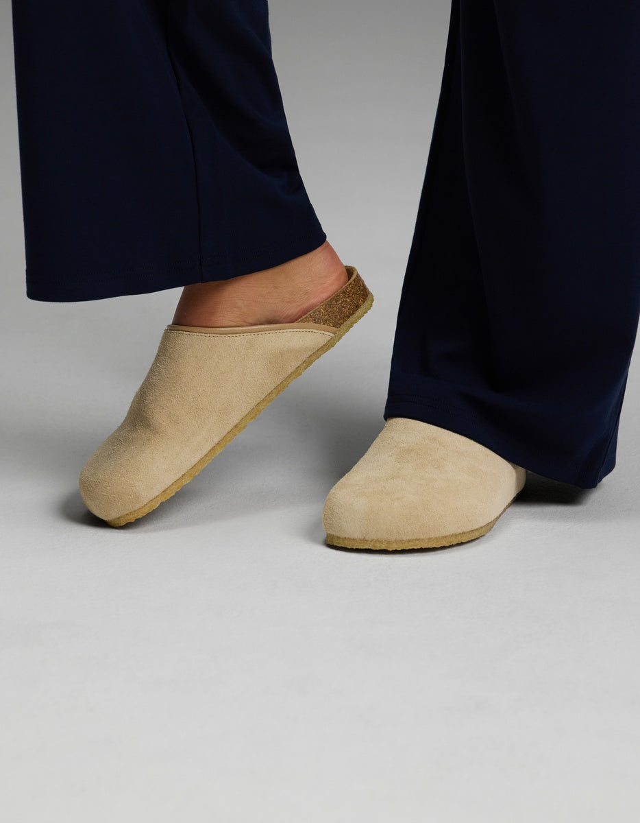 A person wearing navy blue pants and Cozy Earth’s Lakehouse Clog in Suede stands on a light gray surface.