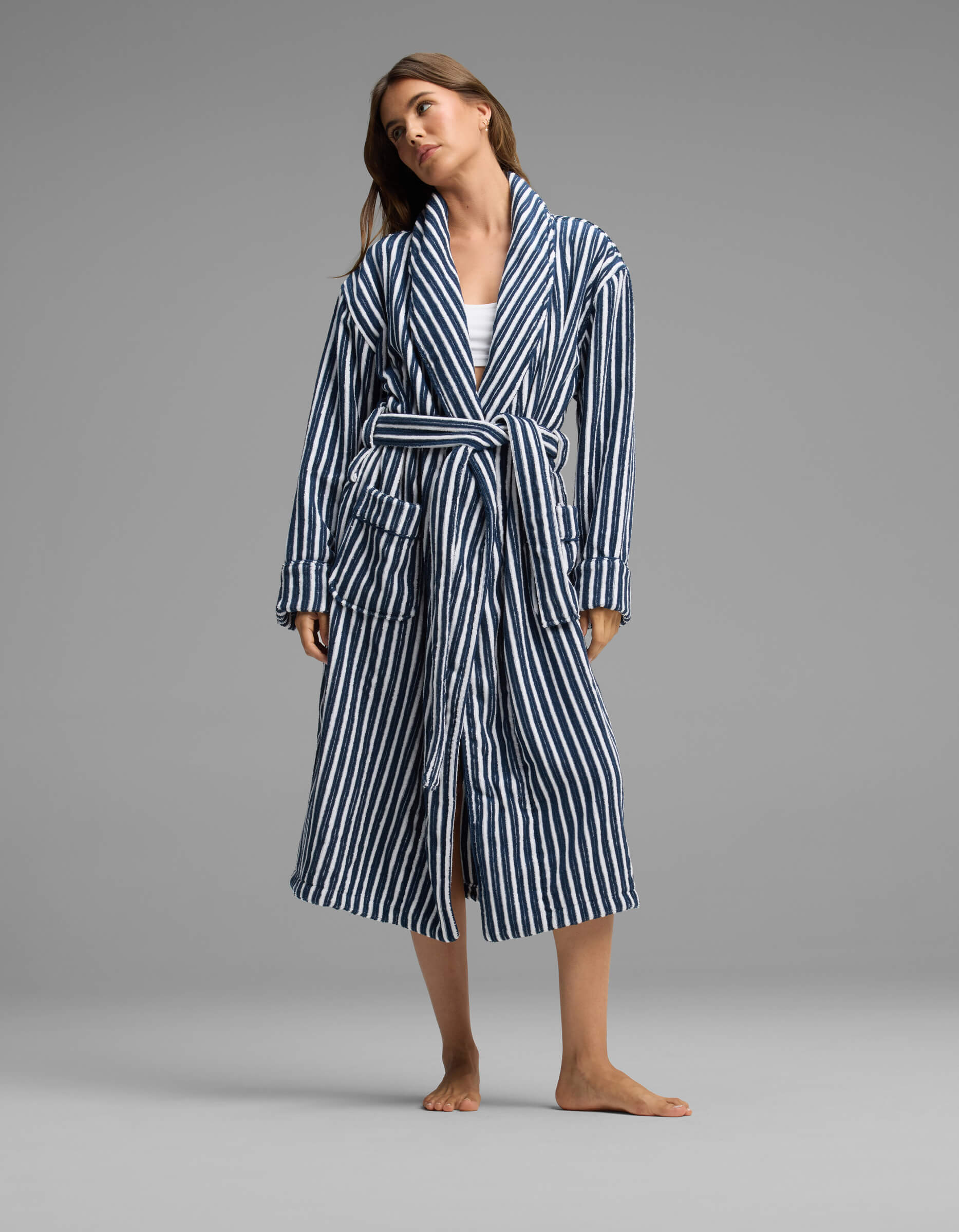 A person stands barefoot against a plain gray background, wearing the Cozy Earth Ribbed Terry Bath Robe—a long, blue-and-white striped robe with a belt—over casual clothes.|Color:Obsidian Stripe