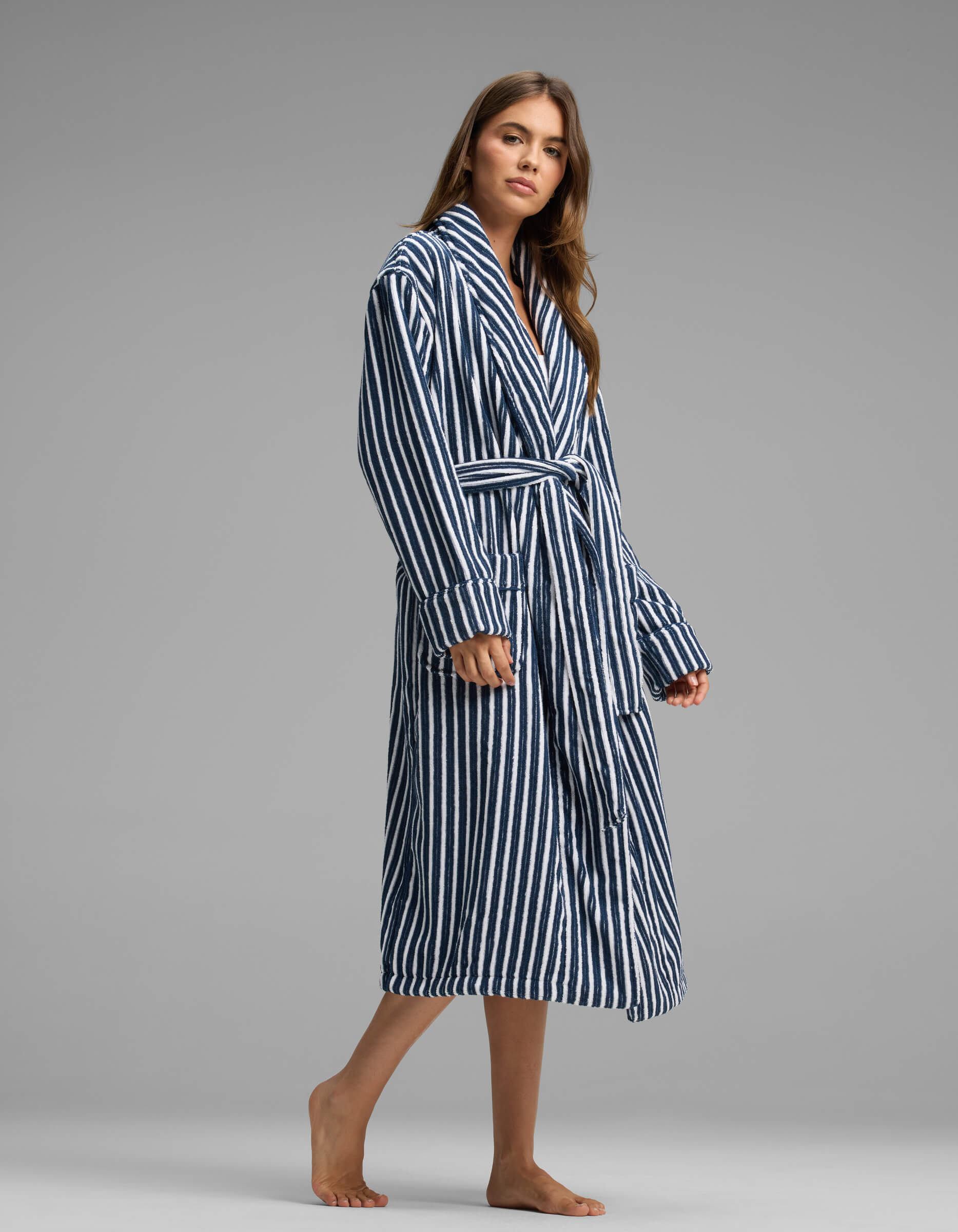 A woman stands barefoot wearing the Cozy Earth Ribbed Terry Bath Robe, featuring a long, blue and white striped design with a belt, against a plain gray background.