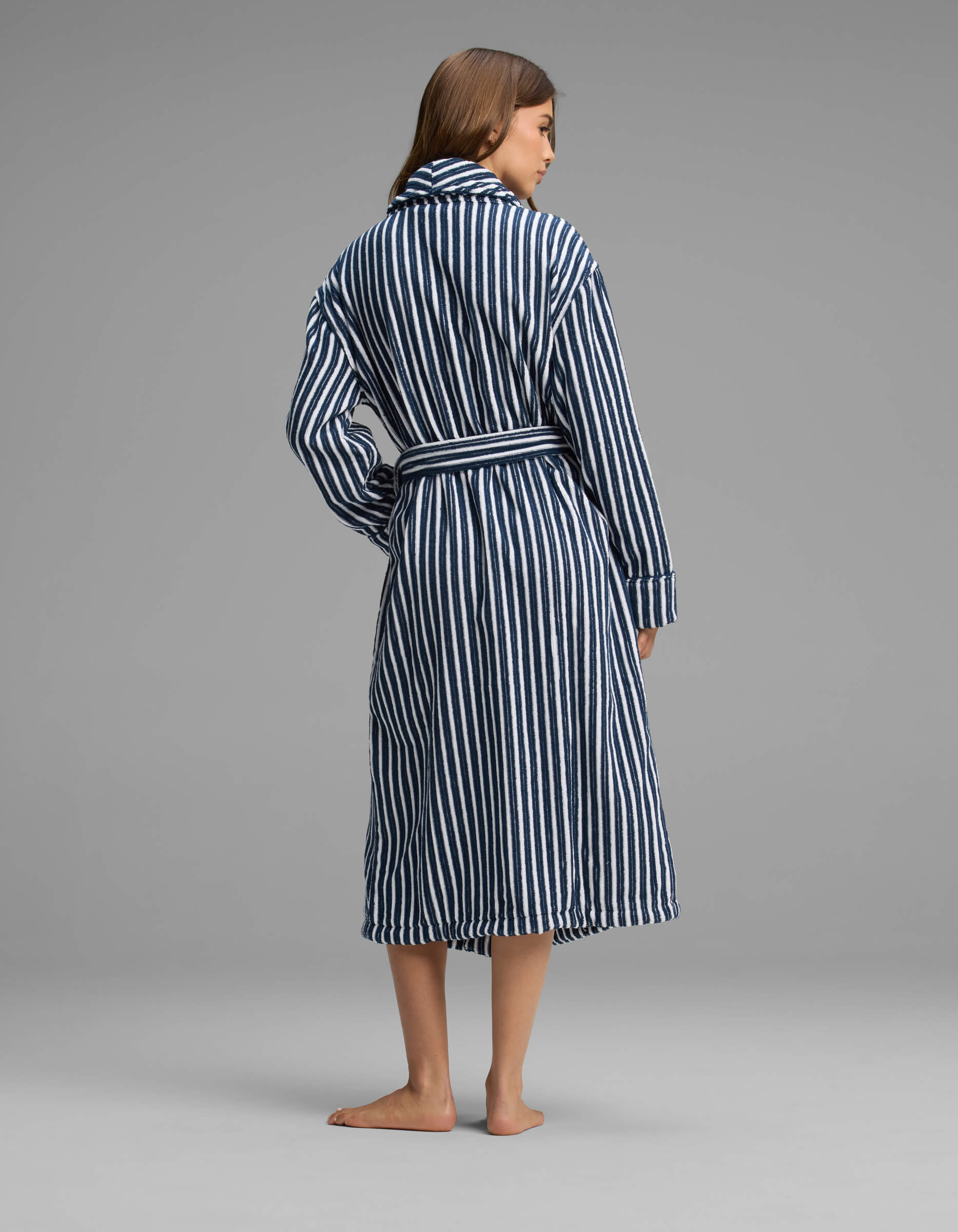 A woman stands barefoot facing away, wearing the Cozy Earth Ribbed Terry Bath Robe in navy and white vertical stripes with a belt tied at the waist, against a plain gray background.