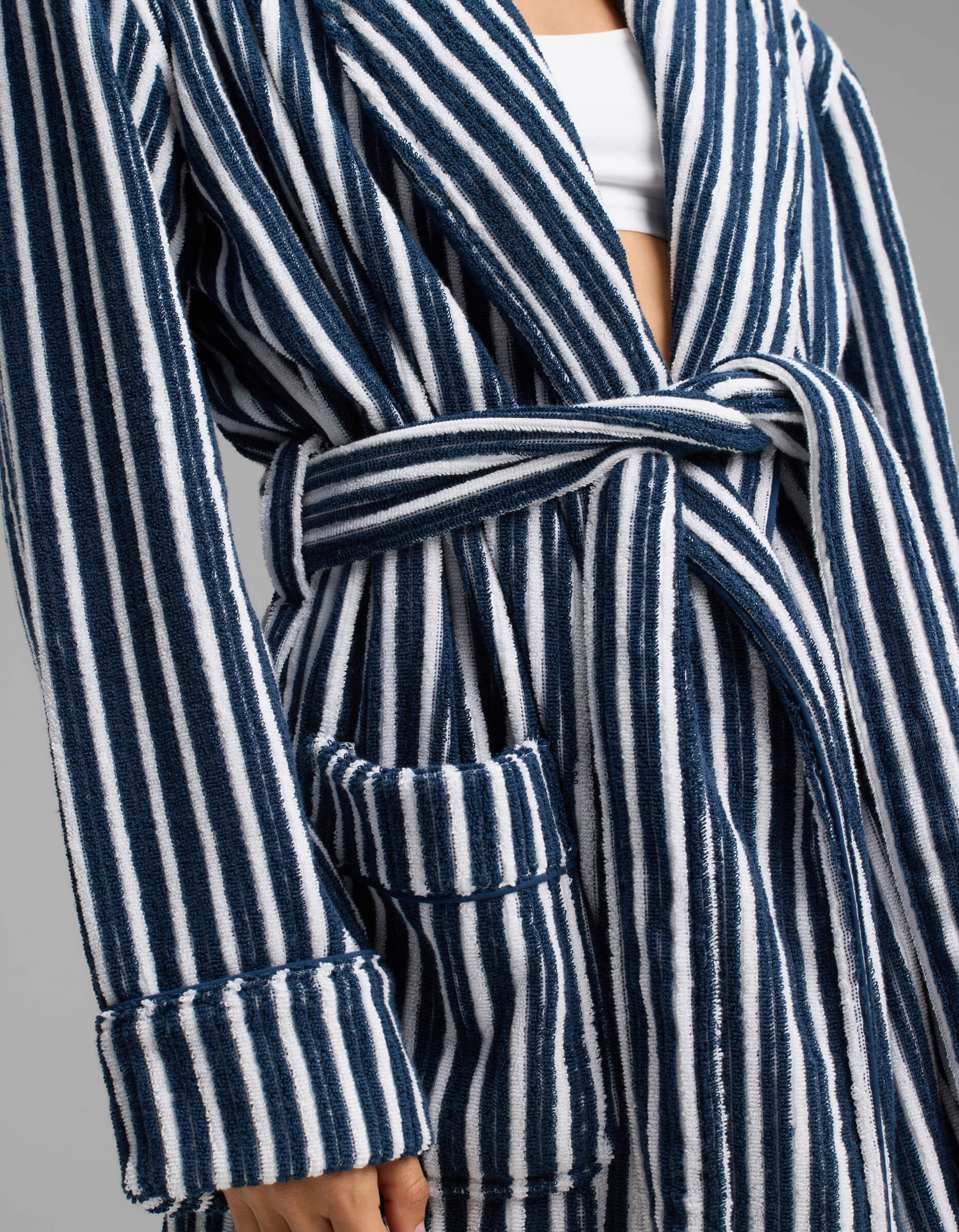 A person wearing the Cozy Earth Ribbed Terry Bath Robe, featuring blue and white vertical stripes, a waist tie belt, and a visible front pocket. The robe is layered over a white top, with only the torso and part of the arm shown.