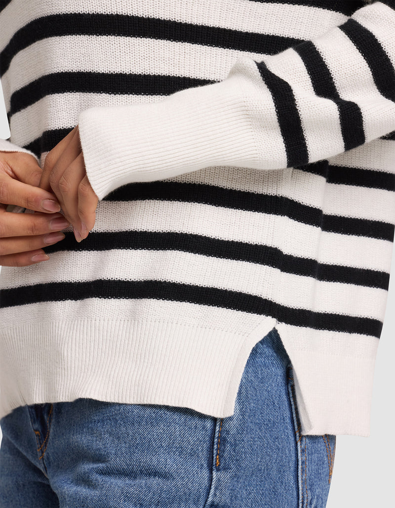 A woman wears the Cozy Earth Women's Rowan Sweater—white with black stripes and a side slit hem—paired with blue jeans, hands clasped near her waist.