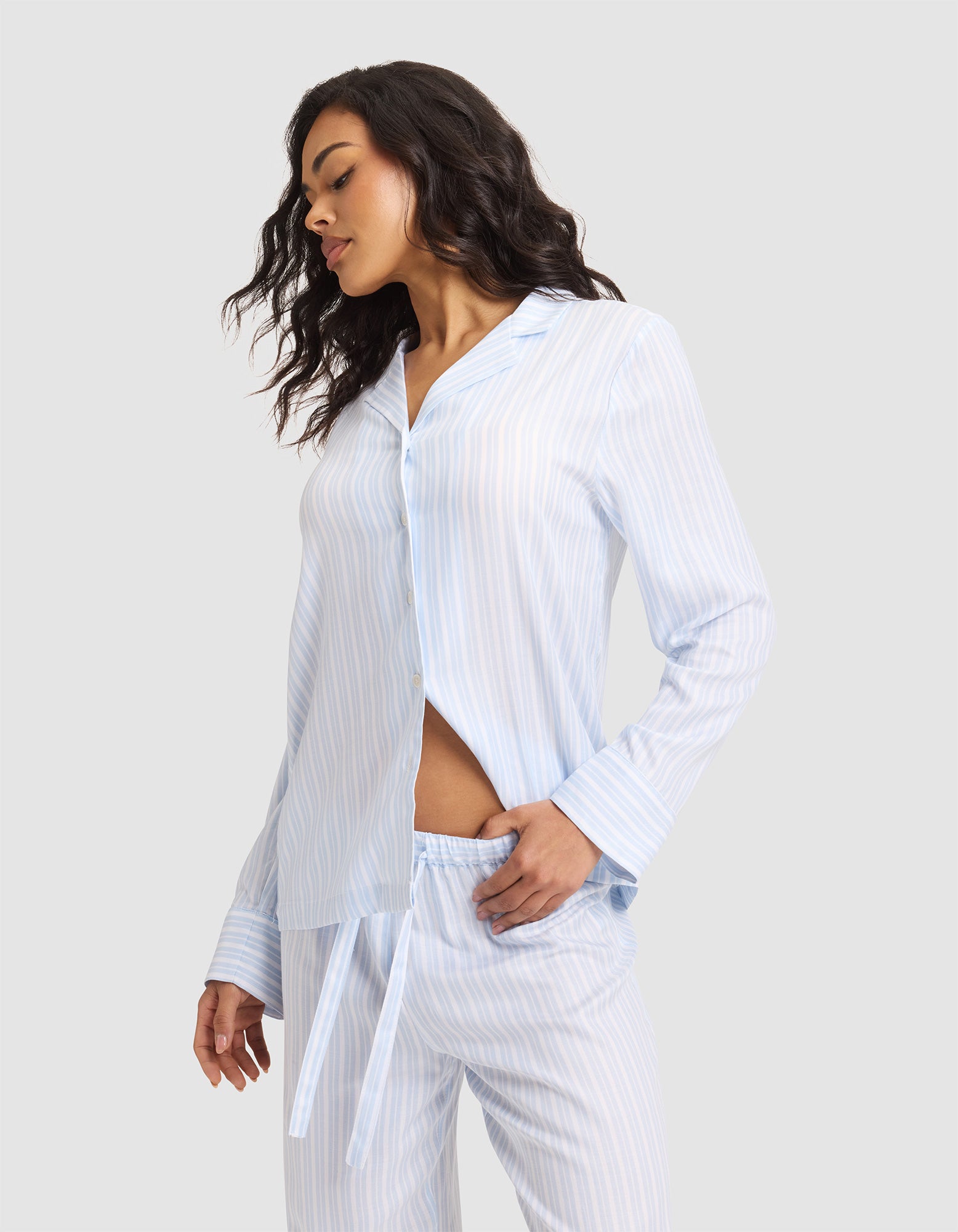 A woman stands sideways in a Cozy Earth Women's Soft Woven Long Sleeve Pajama Top, light blue and white striped, with buttons and matching pants, one hand in her pocket against a plain light background. 