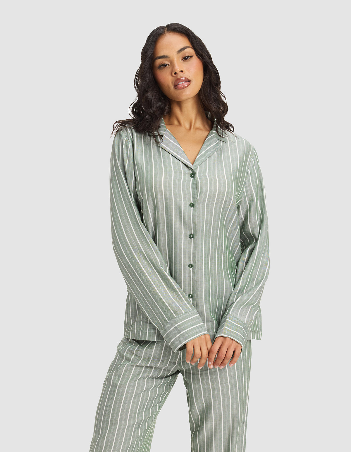 A woman with long, wavy hair models the Cozy Earth Women's Soft Woven Long Sleeve Pajama Top in green and white stripes, standing against a plain light gray background with her hands gently clasped in front.|Color:Striped Jasper