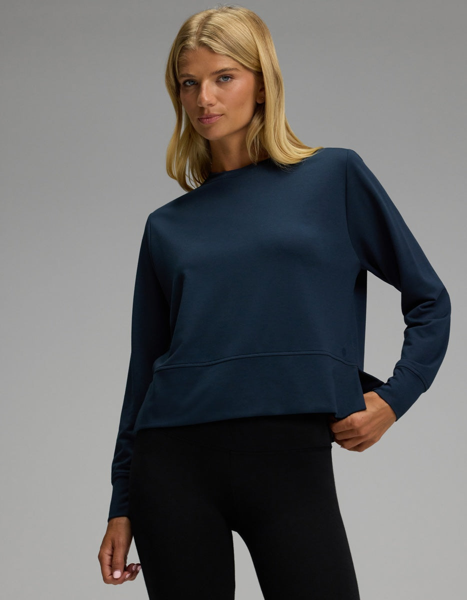 A woman models the Cozy Earth Women’s StudioLite Crewneck in dark blue, paired with black leggings, as she poses with one hand on her hip against a plain gray background.|Color:Eclipse