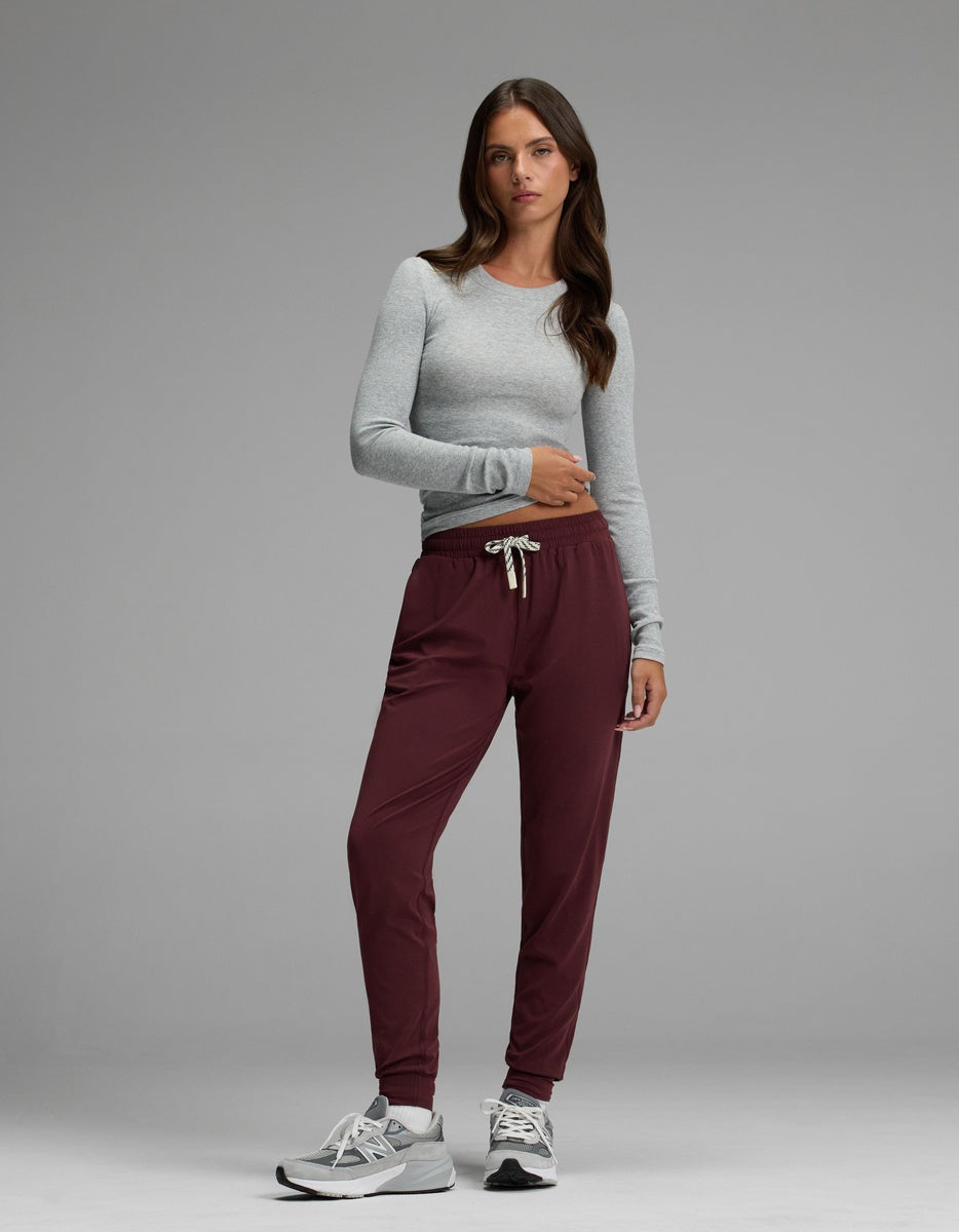 A woman with long brown hair and a neutral expression stands against a gray background wearing the Cozy Earth Women’s Studio Jogger paired with a light gray long-sleeve shirt and gray sneakers.