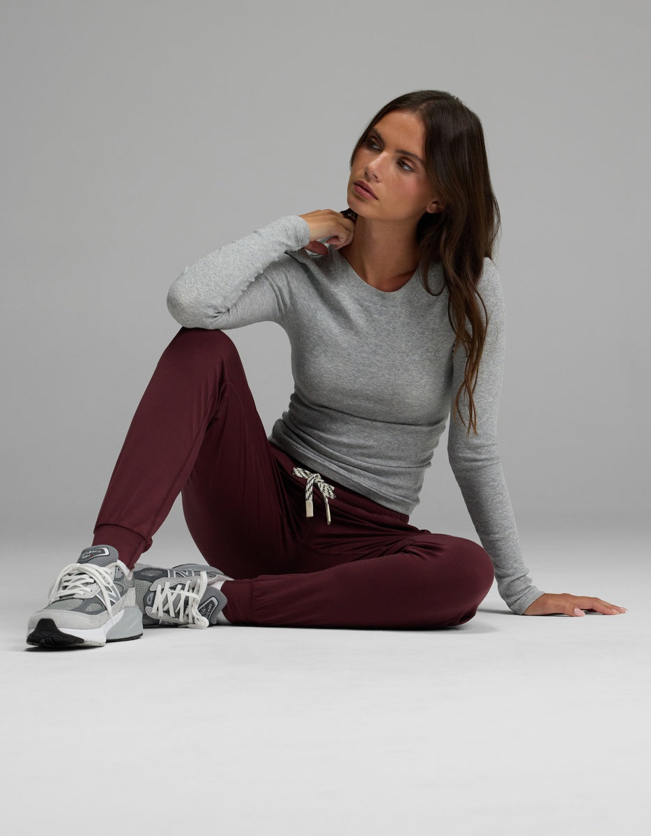A woman wearing Cozy Earth’s Women’s Studio Jogger in maroon and a gray long-sleeve shirt sits on the floor with one knee up, resting her arm on it and gazing to the side. She finishes her look with gray sneakers against a plain light background.