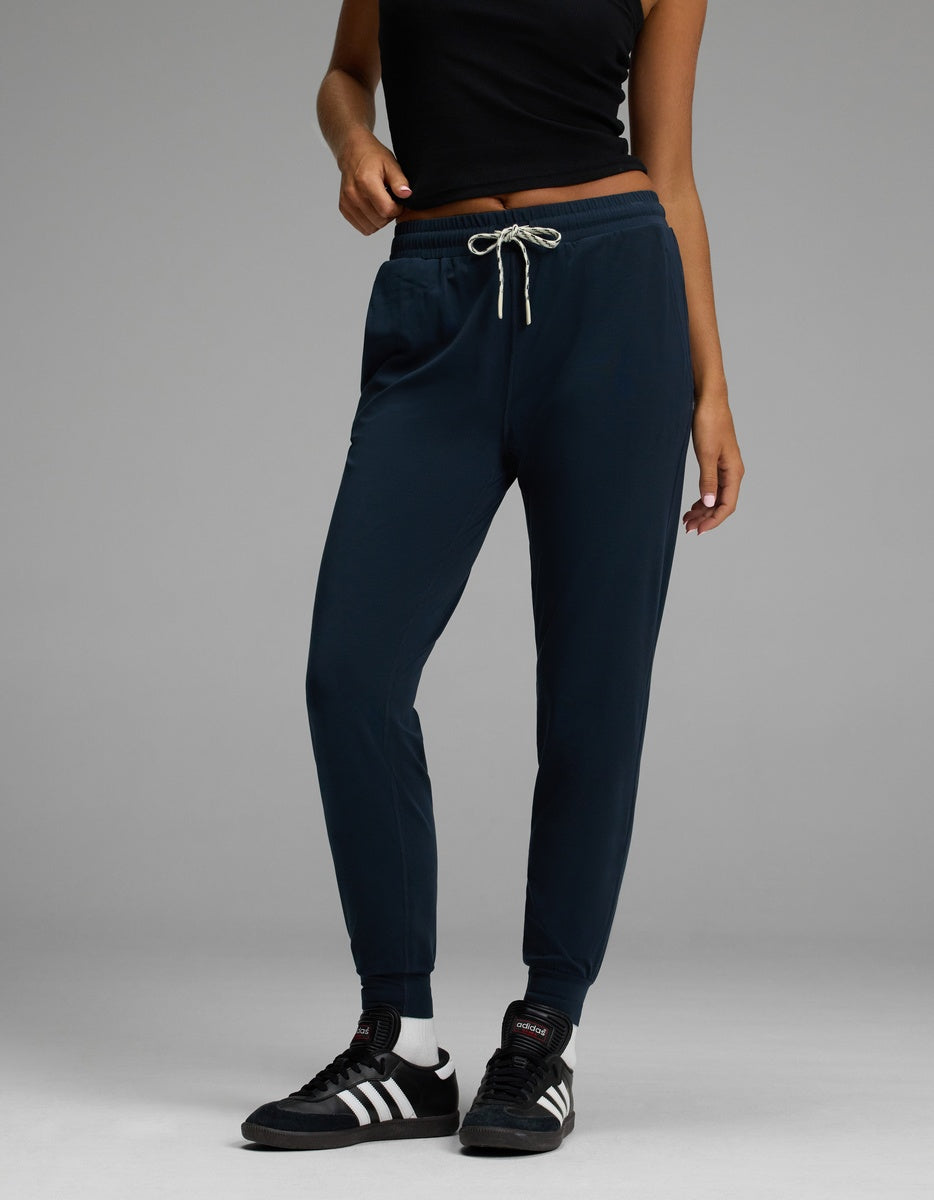 Wearing the Cozy Earth Women’s Studio Jogger in navy, a person stands against a plain gray background, styled with a black sleeveless top, white socks, and black Adidas sneakers.