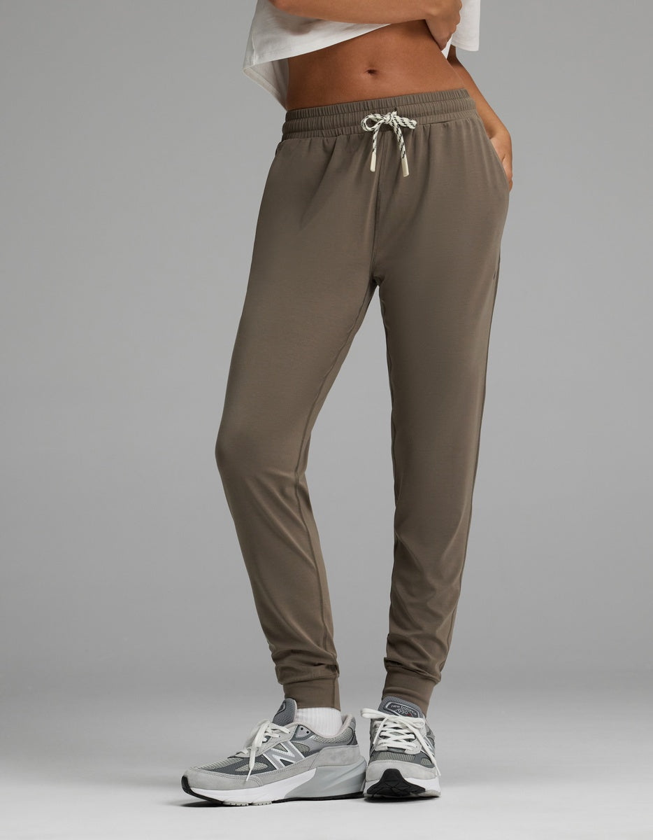 A person is wearing Cozy Earth’s Women’s Studio Jogger in taupe featuring a drawstring waistband, paired with gray sneakers and a white cropped top. The image has a plain gray background and highlights the lower body and legs.