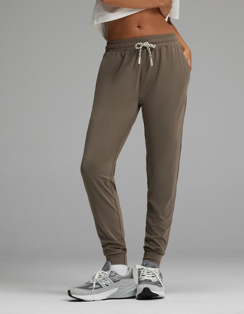 A person is wearing Cozy Earth’s Women’s Studio Jogger in taupe featuring a drawstring waistband, paired with gray sneakers and a white cropped top. The image has a plain gray background and highlights the lower body and legs.